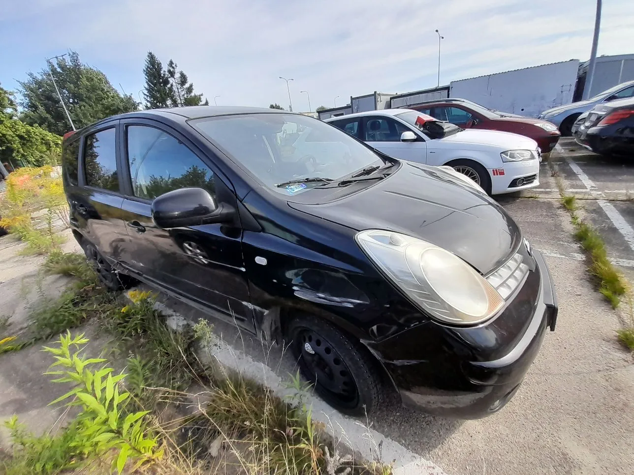 Czarny samochód osobowy typu hatchback zaparkowany na betonowym placu z kępami traw i chwastów. Pojazd jest brudny i posiada widoczne uszkodzenia karoserii na przednim zderzaku oraz boku. W tle widoczne są inne zaparkowane samochody w kolorach białym i czerwonym oraz fragmenty budynków przemysłowych pod jasnym, lekko zachmurzonym niebem.