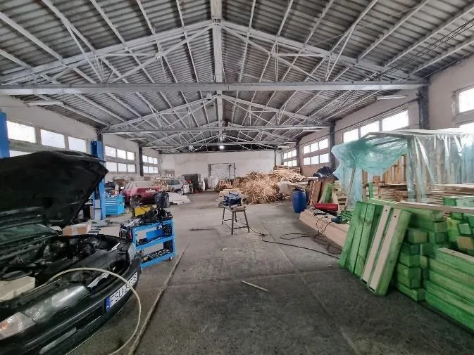 Wnętrze przestronnej hali warsztatowej z metalowym dachem i belkami. Po lewej stronie samochód z otwartą maską, widoczna tablica rejestracyjna z częściowo czytelnym numerem ESU 0. W tle inne pojazdy (czerwony i biały), niebieskie wózki narzędziowe, stół warsztatowy z narzędziami, sterta słomy, stosy zielonych desek oraz materiały przykryte folią. Podłoga betonowa, wysokie okna po bokach hali.