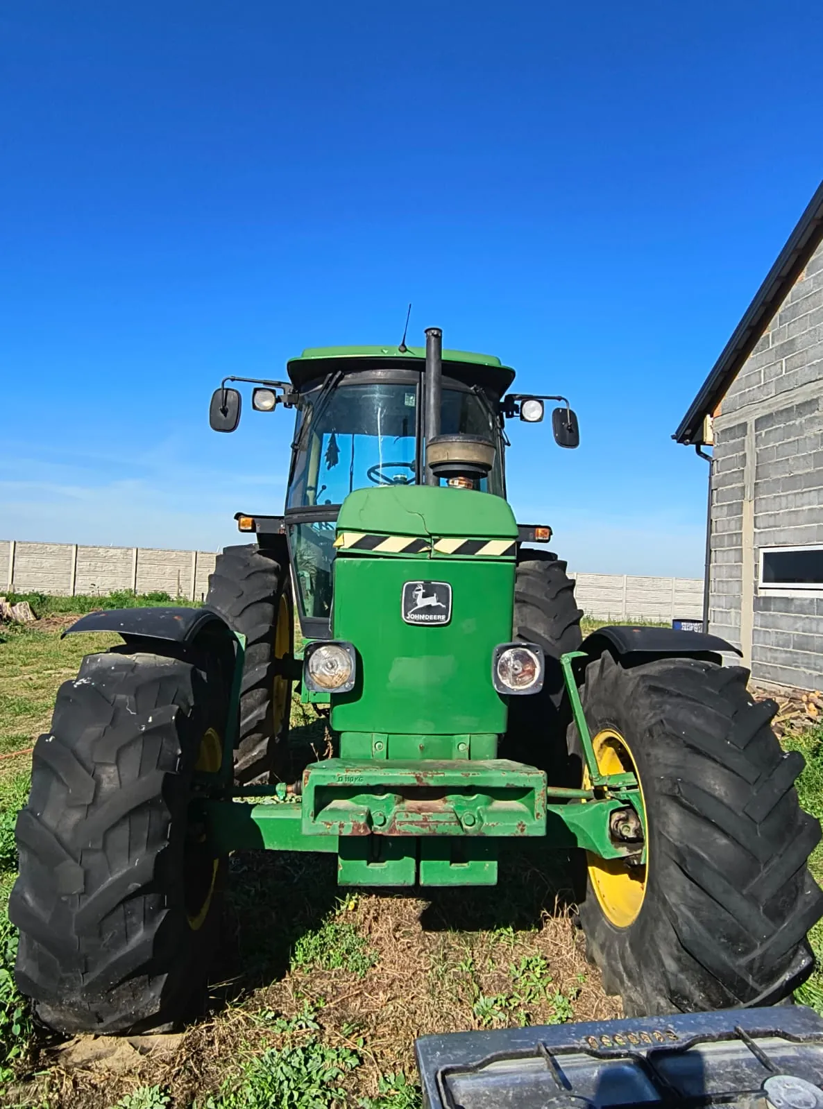 Zielony traktor marki John Deere ustawiony przodem do widza na trawiastym terenie. Maszyna posiada duże, czarne opony z wyraźnym bieżnikiem, kabinę z przeszklonymi szybami i lusterka boczne. Na masce widoczne jest logo John Deere oraz żółto-czarne pasy ostrzegawcze. W tle znajduje się jasny betonowy mur oraz fragment szarego budynku po prawej stronie pod bezchmurnym, niebieskim niebem.