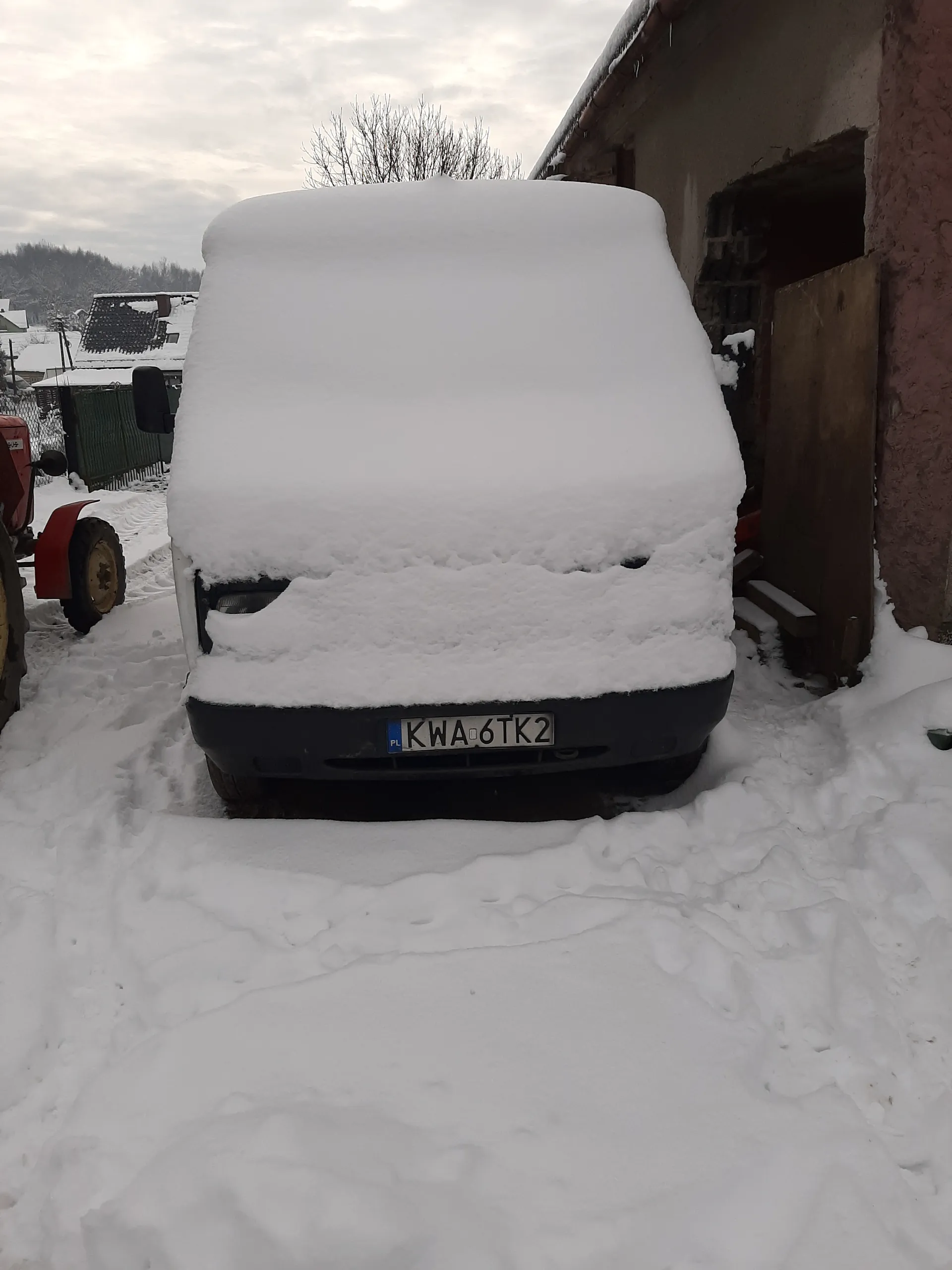 Czarny samochód całkowicie pokryty grubą warstwą śniegu z widoczną tablicą rejestracyjną KWA6TK2. W tle budynek z czerwoną ścianą, drzewa bez liści, śnieżna droga oraz pochmurne niebo. Na pierwszym planie czerwony pojazd rolniczy z kołem i ślady opon w śniegu.
