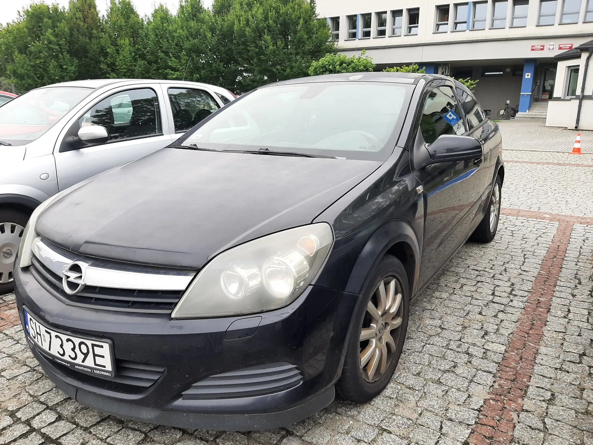 Czarny samochód marki Opel zaparkowany na kostce brukowej. Widoczna jest przednia część pojazdu z tablicą rejestracyjną o numerze SH 7339E. Obok stoi srebrny samochód. W tle widać zielone drzewa oraz nowoczesny, jasny budynek biurowy z wieloma oknami.