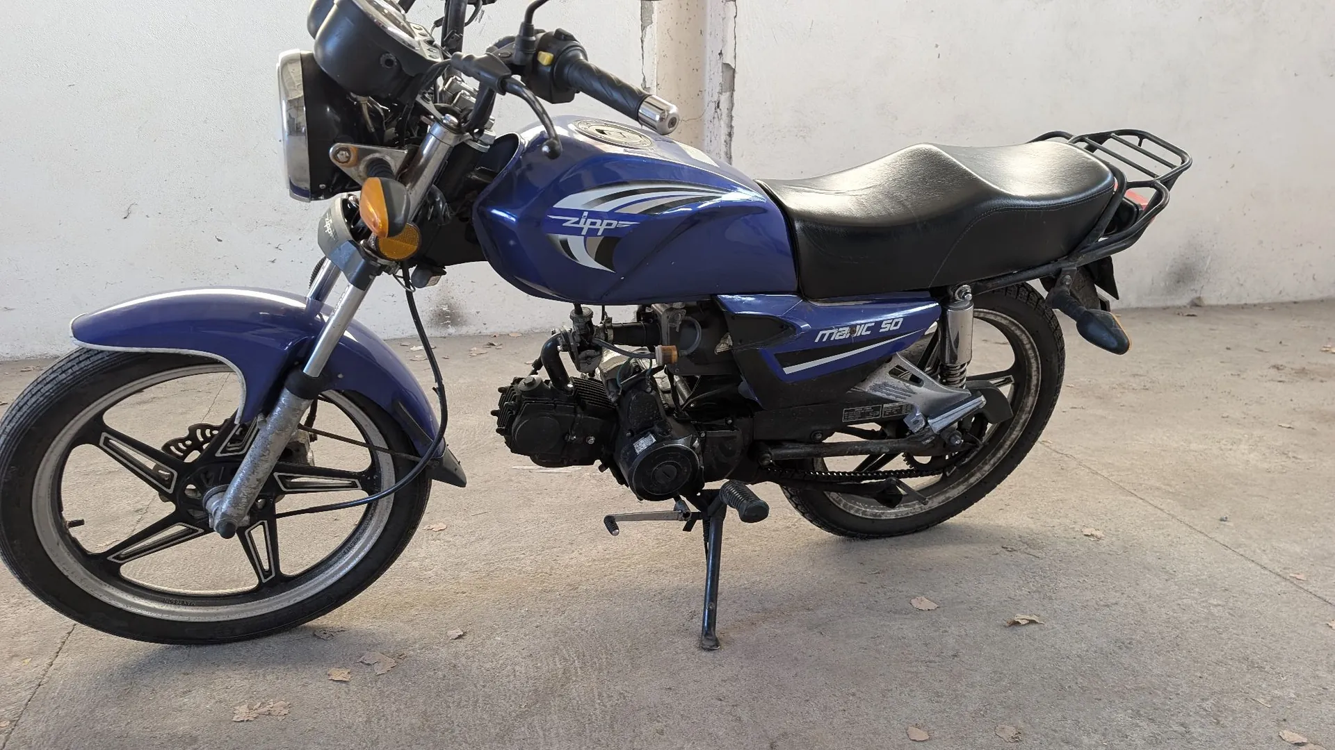 Niebieski motocykl marki Zipp, model Magic 50, zaparkowany na betonowym podłożu. Pojazd posiada czarne siedzenie, tylny bagażnik oraz czarne felgi. Na baku widnieje biały napis 'zipp', a na bocznej owiewce napis 'MAGIC 50'. Motocykl stoi na bocznej stopce.