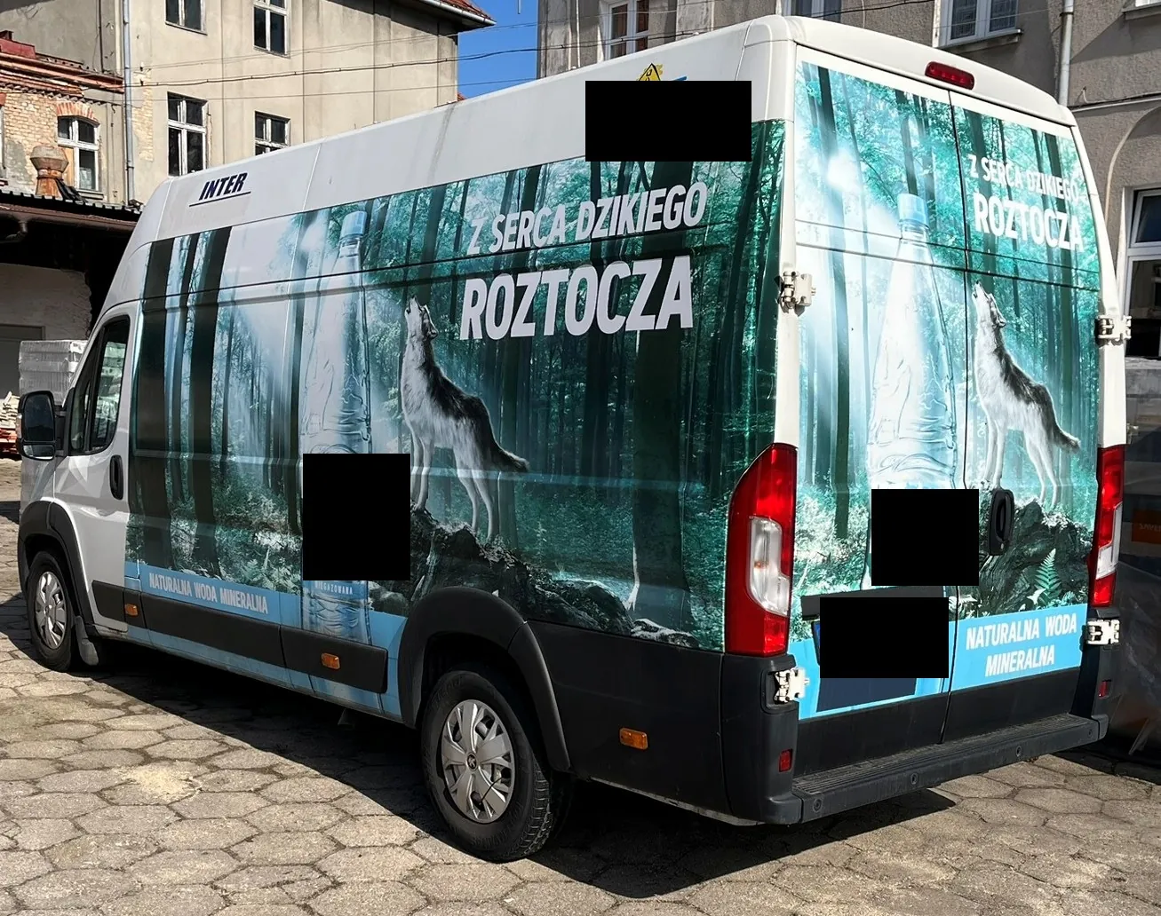 Biały samochód dostawczy z reklamą wody mineralnej na bokach i tylnych drzwiach. Grafika przedstawia gęsty, mglisty las w odcieniach zieleni i błękitu, sylwetkę wilka stojącego na skale oraz dużą butelkę wody. Na karoserii widnieje napis: 'Z SERCA DZIKIEGO ROZTOCZA' oraz mniejszy napis na dole: 'NATURALNA WODA MINERALNA'. W lewym górnym rogu znajduje się logo 'INTER'. Część napisów i elementów graficznych jest zakryta czarnymi prostokątami.