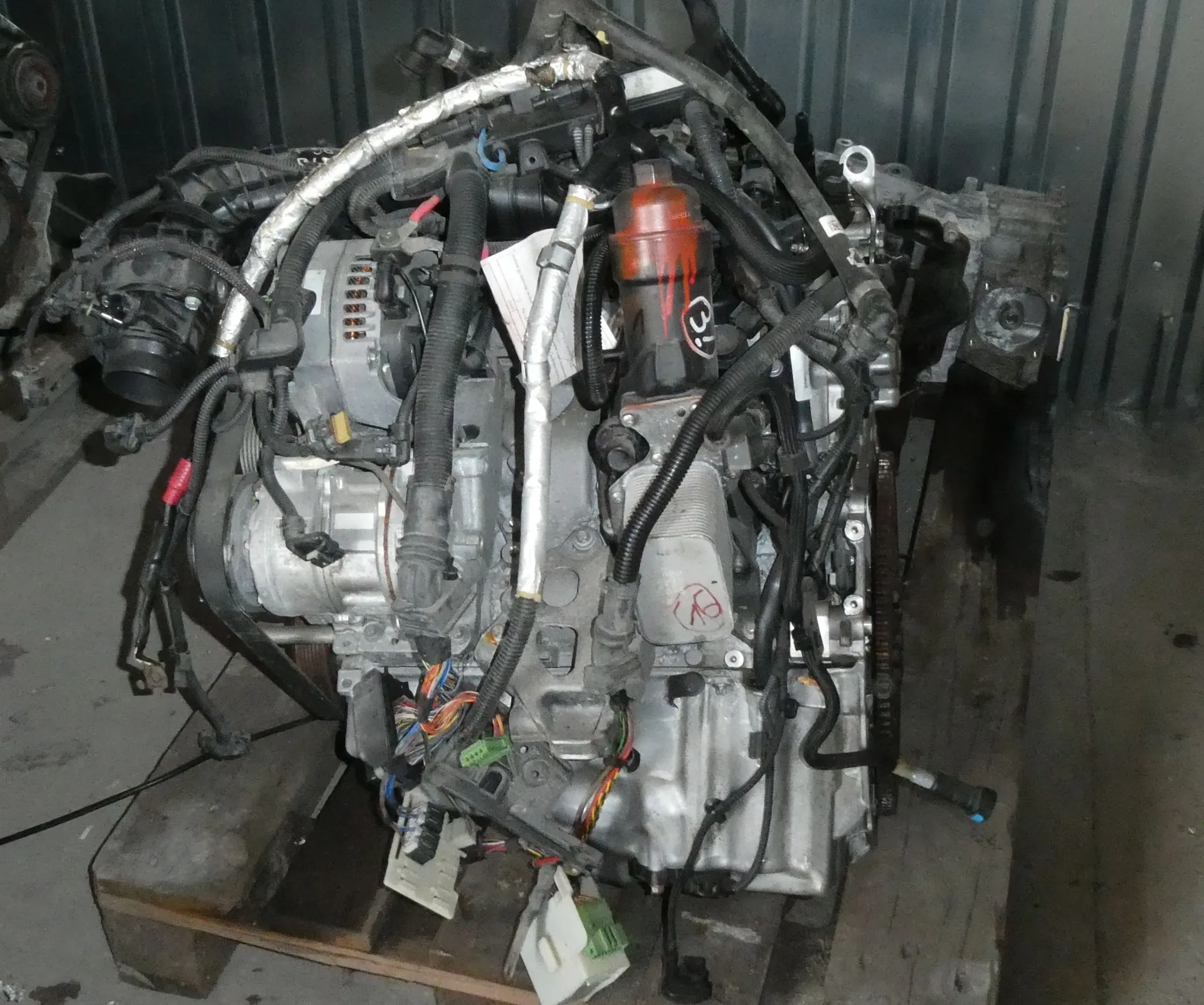 Używany silnik spalinowy z kompletną wiązką elektryczną i osłonami, umieszczony na drewnianej palecie. Widoczne są liczne przewody, rurki, aluminiowe elementy bloku silnika oraz pomarańczowy filtr oleju.