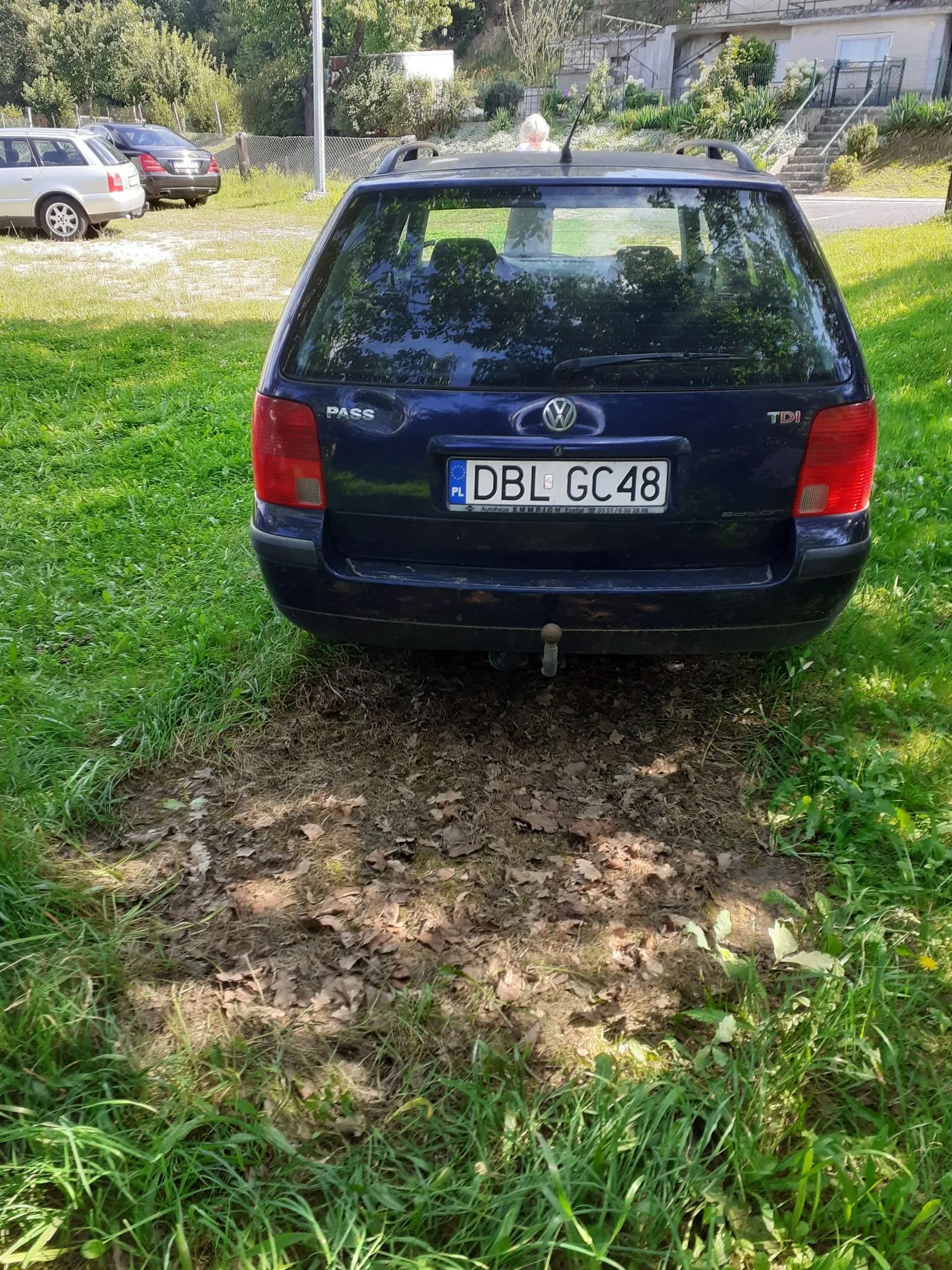 Tylny widok ciemnoniebieskiego samochodu marki Volkswagen Passat TDI zaparkowanego na trawiastym terenie. Samochód ma polską tablicę rejestracyjną o numerze DBL GC48. Przed pojazdem znajduje się obszar z błotem i suchymi liśćmi. W tle widoczne są inne zaparkowane samochody, zieleń oraz fragmenty budynków.