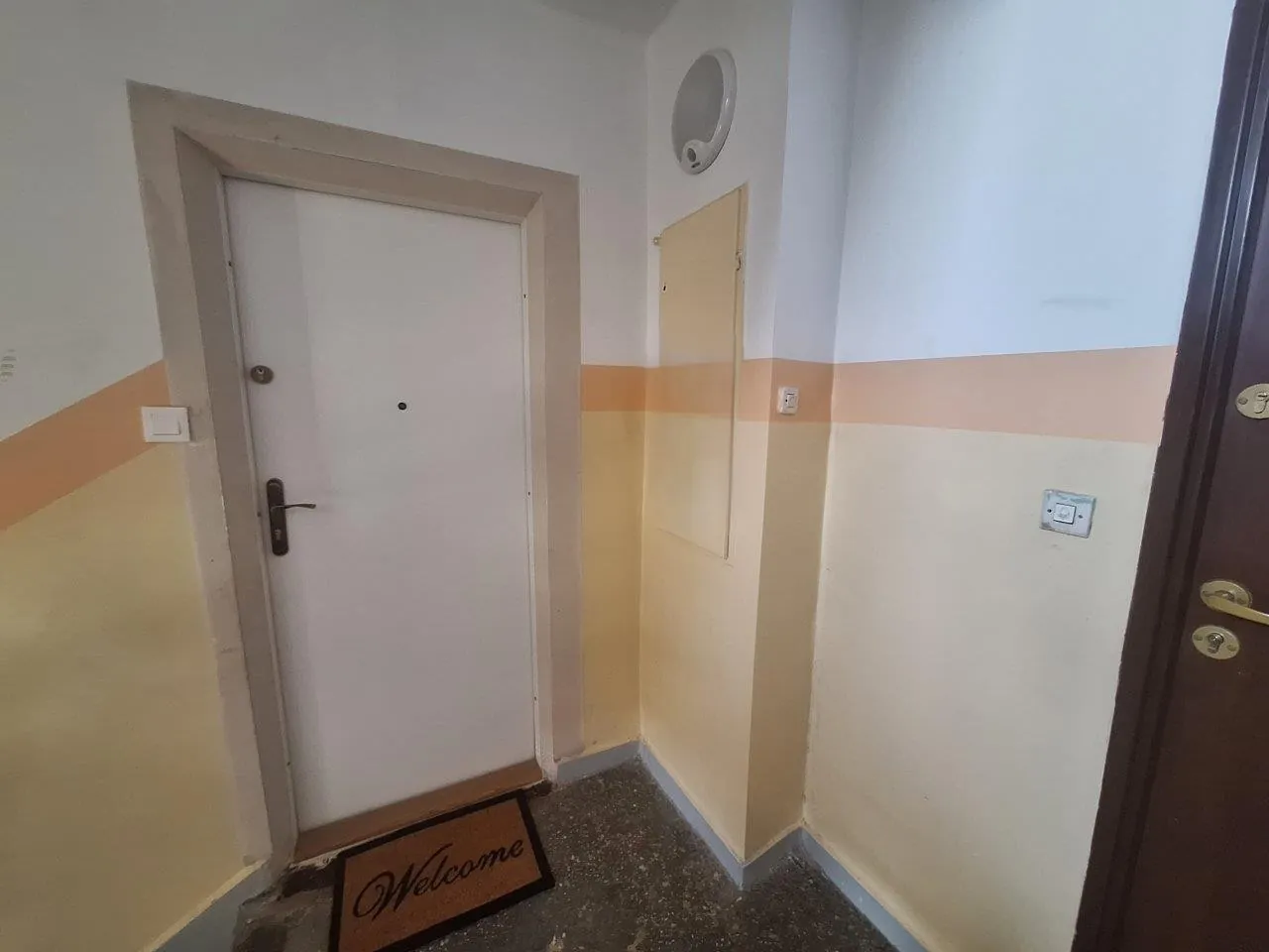 Wnętrze przedpokoju z białymi drzwiami w brązowej ramie. Na podłodze przed drzwiami leży ciemna wycieraczka z napisem 'Welcome'. Ściany są pomalowane w dwóch kolorach: białe u góry i żółte u dołu, rozdzielone pomarańczowym pasem. Po prawej stronie widoczna jest część ciemnobrązowych drzwi oraz gniazdko elektryczne na ścianie.