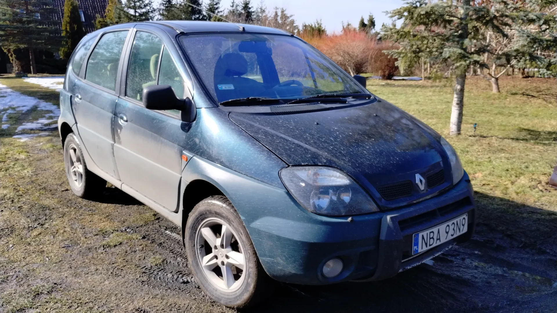Niebieski samochód osobowy marki Renault z tablicą rejestracyjną NBA 93N9 (PL) zaparkowany na trawiastym i błotnistym terenie. W tle drzewa, krzewy oraz resztki śniegu. Samochód ma zabrudzoną karoserię i srebrne felgi.
