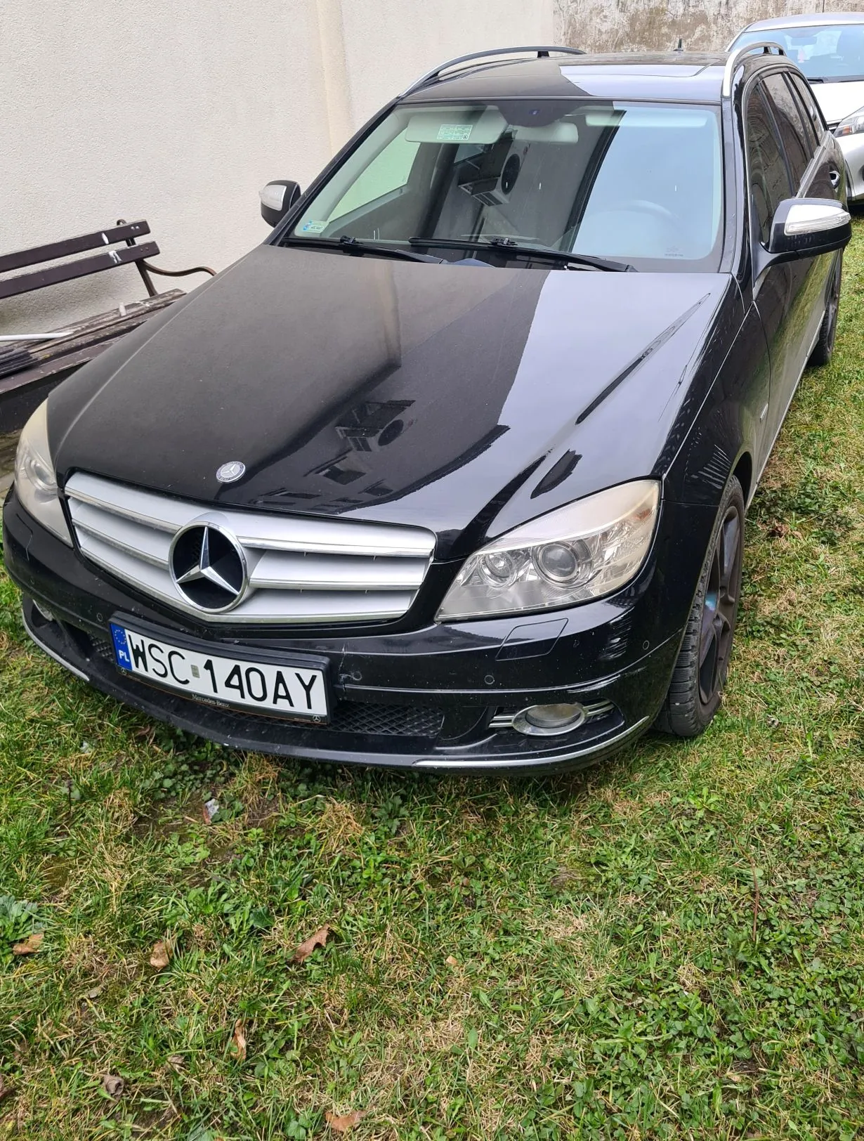 Czarny samochód osobowy typu kombi marki Mercedes-Benz zaparkowany na trawie. Widoczna tablica rejestracyjna z polskim kodem "PL" i numerem "WSC 140AY". W tle biała ściana budynku, ławka oraz fragment innego samochodu.