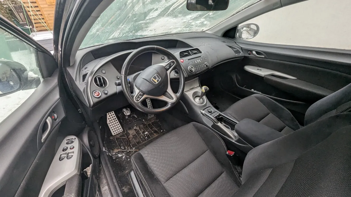 Wnętrze samochodu marki Honda z widoczną kierownicą z logo, czarnymi siedzeniami, brudną podłogą z resztkami śniegu i otwartą drzwiami po stronie kierowcy. Na desce rozdzielczej znajdują się przyciski, radio oraz klimatyzacja. Przez przednią szybę widoczny jest zasneżony teren i drewniana konstrukcja. W samochodzie widoczne są pedały, dźwignia zmiany biegów oraz lusterko wsteczne.