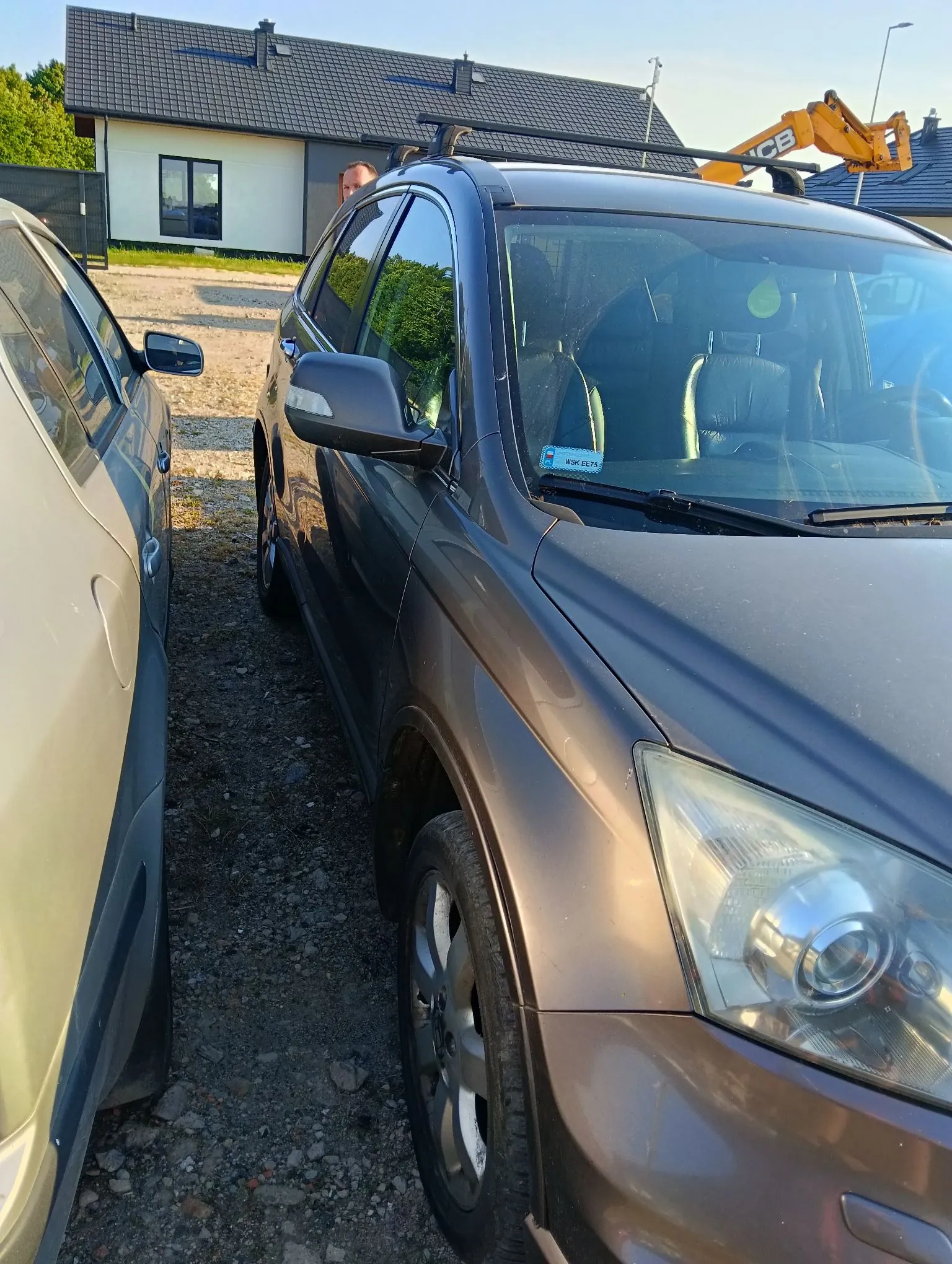 Zdjęcie przedstawiające przód i bok ciemnobrązowego samochodu typu SUV zaparkowanego na żwirowym podjeździe obok innego, jasnego samochodu. W tle widoczny jest biały dom z ciemnym dachem oraz żółta koparka. Na przedniej szybie ciemnego auta znajduje się niebieska naklejka z tekstem 'RVK 5679'.