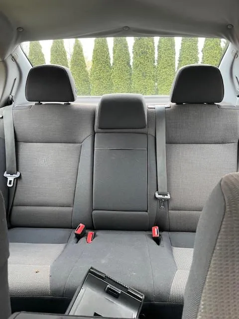 Wnętrze samochodu widziane od tyłu przednich siedzeń. Trzy tylne siedzenia z szarymi poduszkami i czarnymi zagłówkami. Widoczne pasy bezpieczeństwa z czerwonymi zapięciami. Przez tylne okno widać rzęd wysokich zielonych drzew. Na środkowym siedzeniu leży czarny przedmiot przypominający etui lub tablet.