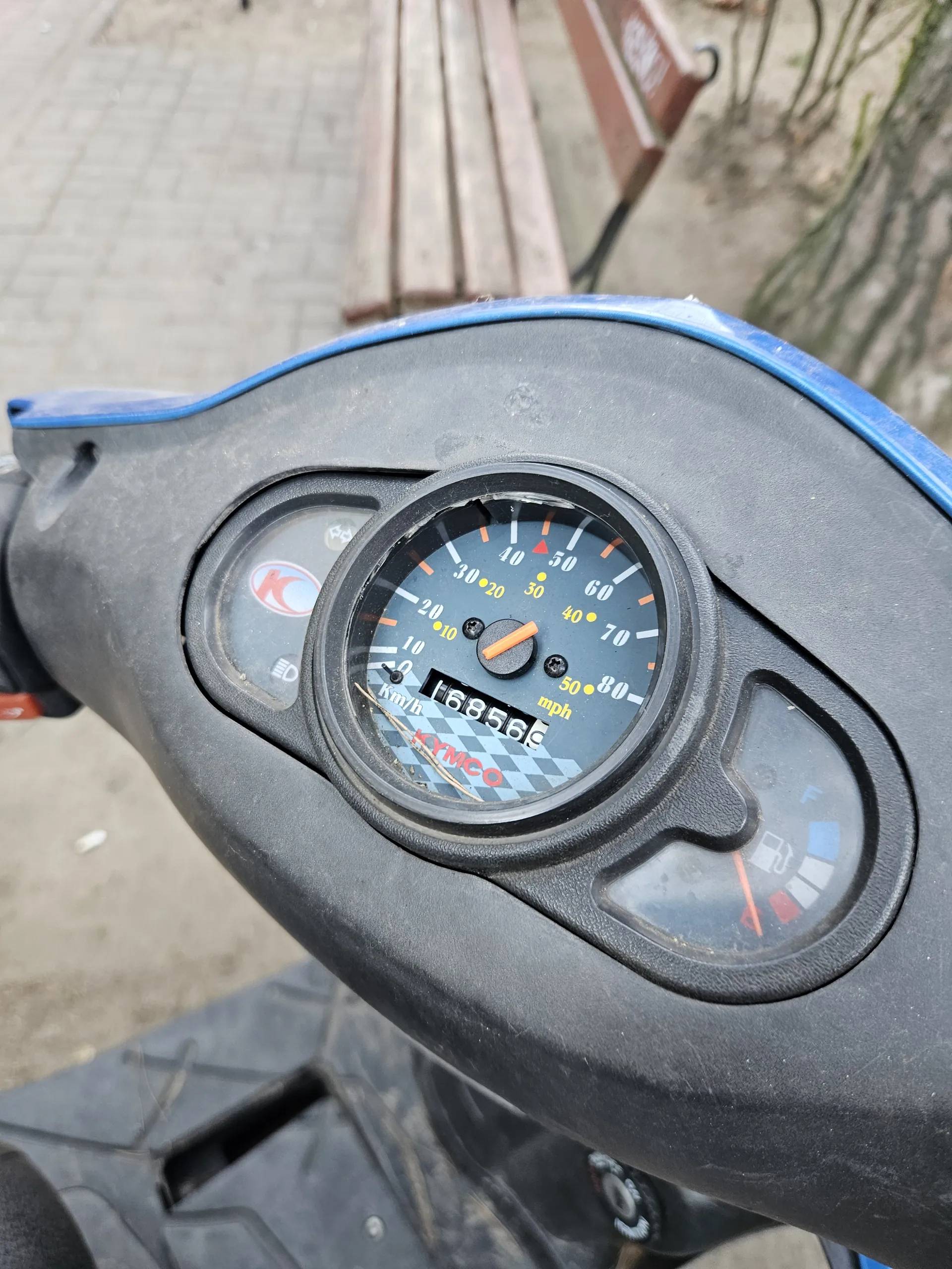 Zbliżenie na desce rozdzielczej skutera KYMCO. W centrum okrągły wskaźnik prędkości z zakresem od 0 do 80 km/h, cyfrowy licznik przebiegu pokazujący 16856 km. Powyżej wskaźnik paliwa z czerwoną igłą wskazującą na niski poziom. W tle widoczna ławka i drzewa.