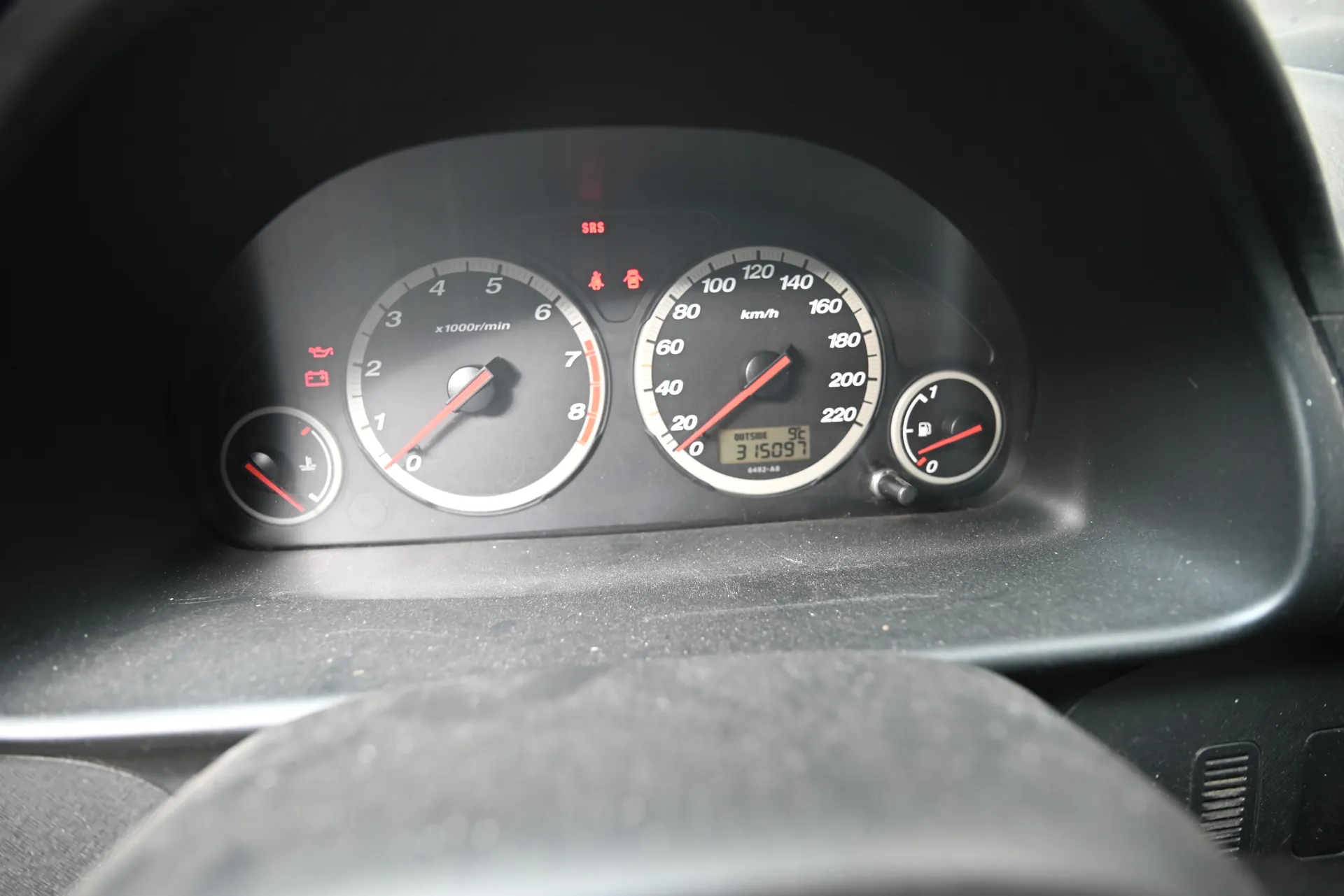 Zbliżenie na deski rozdzielcze samochodu z czterema wskaźnikami. Po lewej stronie wskaźnik temperatury chłodniczki i wskaźnik obrotomieru (0-8 x1000r/min) z czerwoną igłą na 0. W centrum wskaźnik prędkości (0-220 km/h) z czerwoną igłą na 0 i wyświetlaczem przebiegu 315097. Po prawej stronie wskaźnik paliwa z czerwoną igłą blisko 0. Na górze czerwone sygnalizatory: SRS, pas bezpieczeństwa i inny symbol. W dolnej części widoczny fragment kierownicy. Tło czarne, wskaźniki z białymi cyframi i czerwonymi igłami.