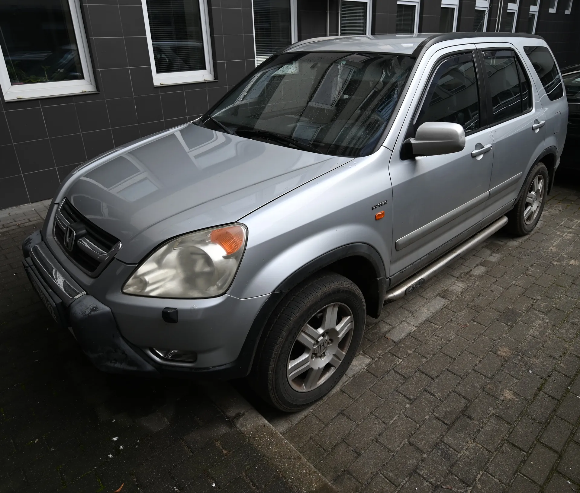 Srebrny SUV marki Honda zaparkowany na brukowanej powierzchni przed budynkiem z ciemnoszarymi płytkami i białymi oknami. Samochód posiada reflektory, lusterka boczne oraz felgi aluminiowe. W tle widoczne są okna budynku i fragment innego pojazdu.