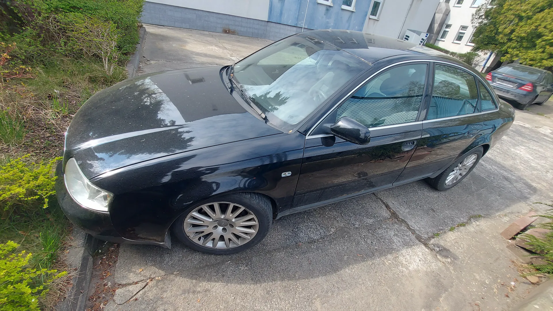 Czarny samochód osobowy typu sedan zaparkowany na betonowej powierzchni. Po lewej stronie obrazu zielone krzewy i trawa, w tle budynek z niebieskimi i białymi ścianami oraz drugi czarny samochód. Samochód posiada srebrne felgi, czarne lusterka boczne i cztery drzwi. Widoczne przednie światło oraz siatka oddzielająca przestrzeń bagażnika od wnętrza pojazdu.