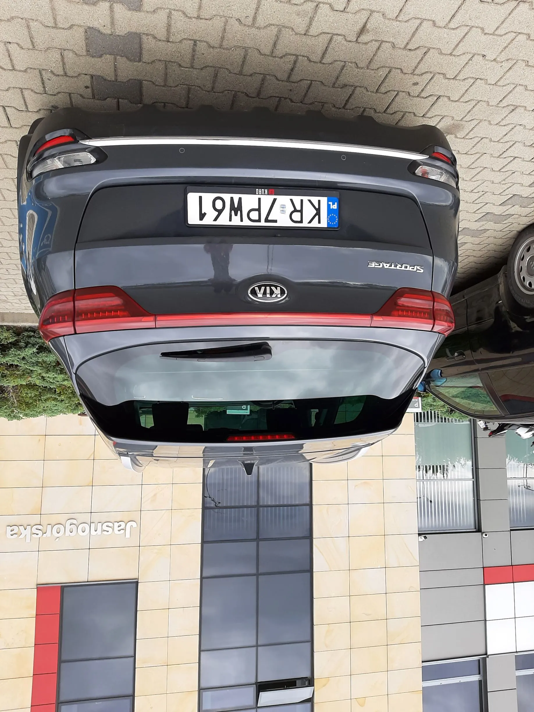 Tył ciemnoszarego samochodu marki Kia Sportage zaparkowanego na kostce brukowej przed budynkiem. Na samochodzie widoczna jest tablica rejestracyjna o numerze KR 7PW61. W tle znajduje się jasna elewacja budynku z napisem „Jasnogórska”.