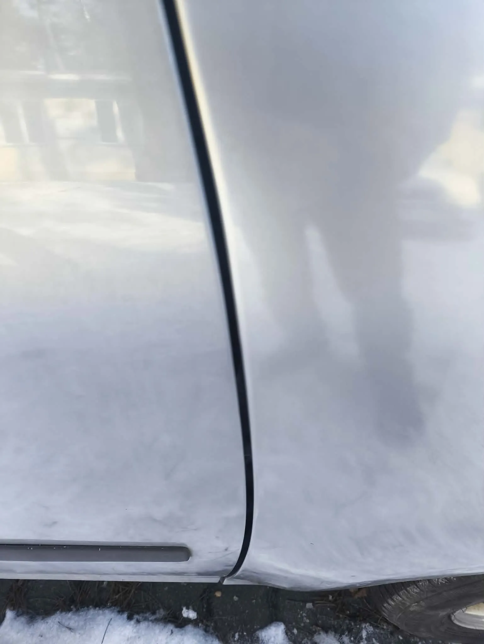 Zbliżenie na bok jasnoszarego samochodu z widoczną czarną linią łączącą panele karoserii. Na błyszczącej powierzchni odbijają się sylwetka budynku i cień stojącej osoby. W dolnej części obrazu widoczny jest fragment asfaltu z resztkami śniegu oraz część opony.