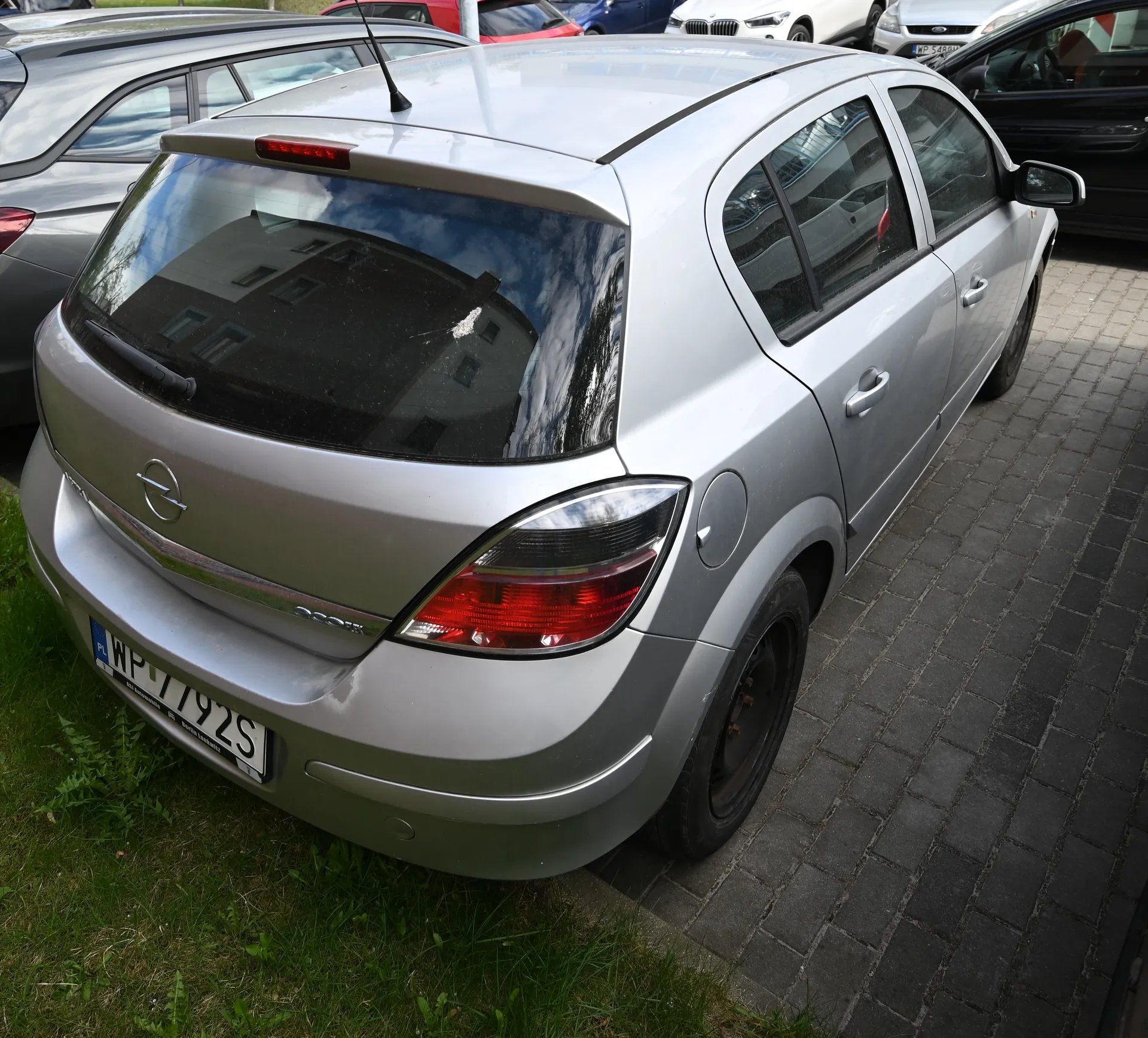 Srebrny samochód Opel Astra z tyłu, z tablicą rejestracyjną WP 7792S, zaparkowany na brukowanej powierzchni obok trawnika. W tle inne samochody (szary, czerwony, biały, niebieski, czarny) oraz odbicie budynku i nieba w tylnej szybie.