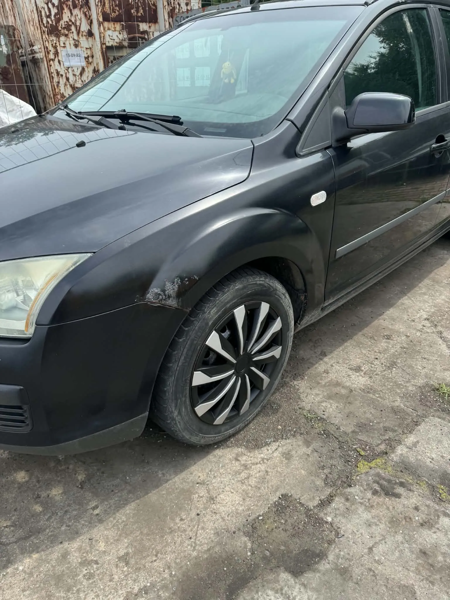 Zbliżenie na przód czarnego samochodu typu minivan. Widoczne jest wgniecenie i uszkodzenie lakieru na przednim lewym błotniku, nad kołem. Samochód posiada wieloramienne felgi aluminiowe w kolorze srebrno-czarnym. W tle widać fragment zardzewiałej, metalowej bramy.