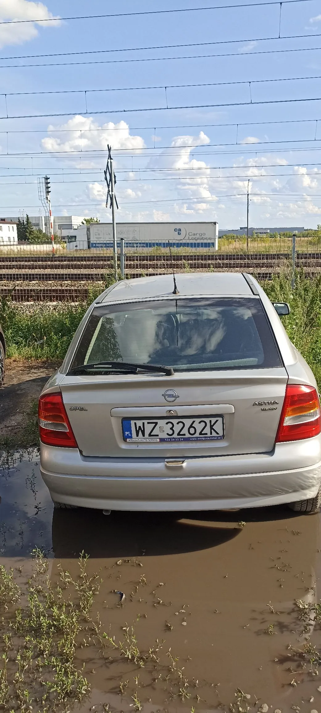 Srebrny samochód marki Opel Astra widziany od tyłu, zaparkowany na błotnistym terenie z kałużami. Na klapie bagażnika znajduje się polska tablica rejestracyjna o numerze WZ 3262K. W tle widoczne są tory kolejowe, ogrodzenie, budynki przemysłowe z napisem 'Cargo Logistics' oraz niebo z białymi chmurami.