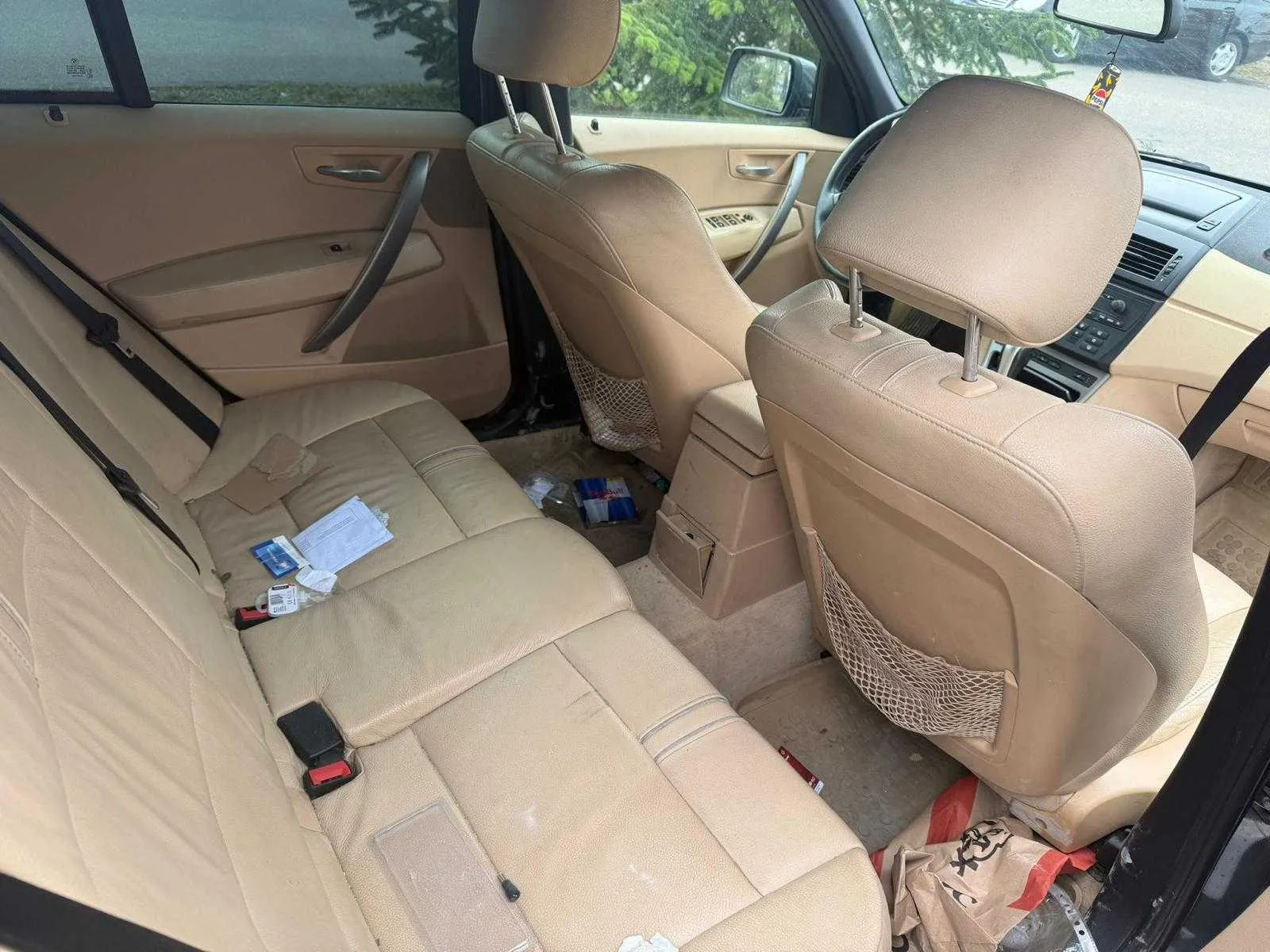 Wnętrze samochodu z widokiem na tylną część kabiny. Beżowe skórzane siedzenia, na tylnym siedzeniu leżą różne przedmioty: biała kartka, niebieska karta, czarno-czerwony przedmiot. Na podłodze widoczna torba KFC, śmieci i siateczka na oparciu przedniego siedzenia. Drzwi samochodu są otwarte, przez okno widać drzewa i inną pojazd. Deska rozdzielcza i kierownica są widoczne, a wnętrze jest w kolorze beżu.