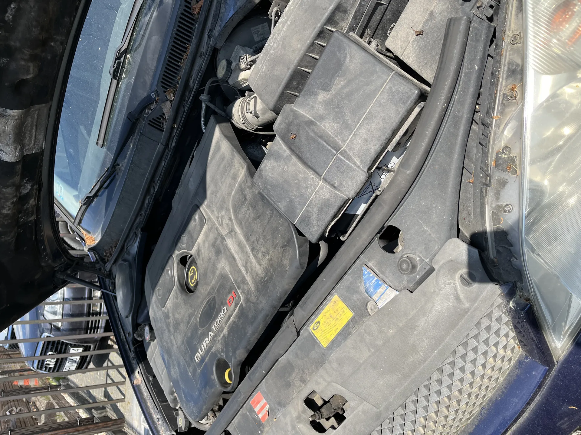 Otwarta kapota samochodu z widocznym silnikiem. Na pokrywie silnika napis 'DURA TORQ DI' oraz żółta etykieta z tekstem. Po prawej stronie widoczny reflektor. W tle drugi samochód i metalowe ogrodzenie. Elementy silnika są zapylone.