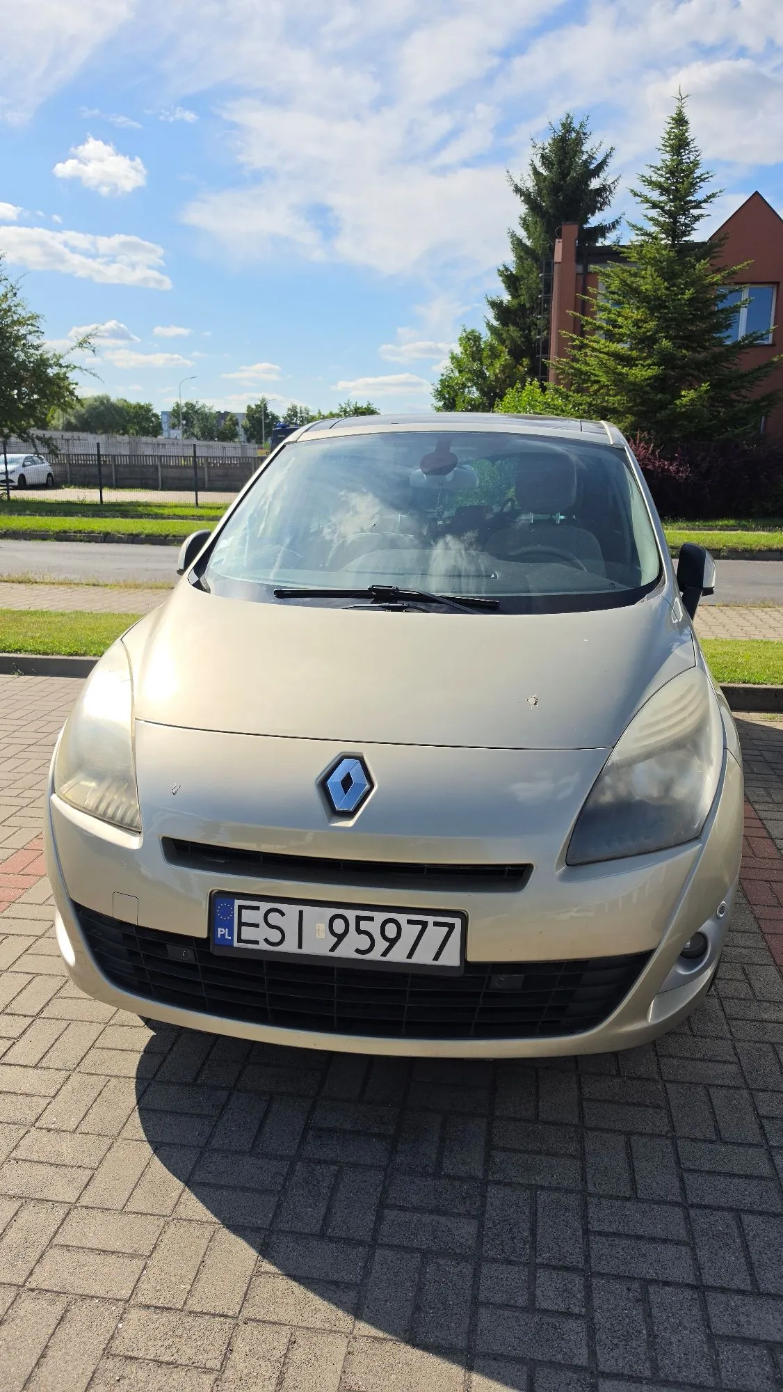 Złoty samochód marki Renault zaparkowany przodem do obiektywu na kostce brukowej. Na samochodzie widoczna jest polska tablica rejestracyjna o numerze ESI 95977. W tle widać zielone drzewa, budynek z brązowej cegły oraz niebo z białymi chmurami.