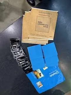 Na ciemnym stole leżą niebieskie folderki z małymi zdjęciami, czarna taśma z białym tekstem oraz stos brązowych kopert z widocznym napisem 'The New York Times'. W prawym dolnym rogu widoczna jest część buta.