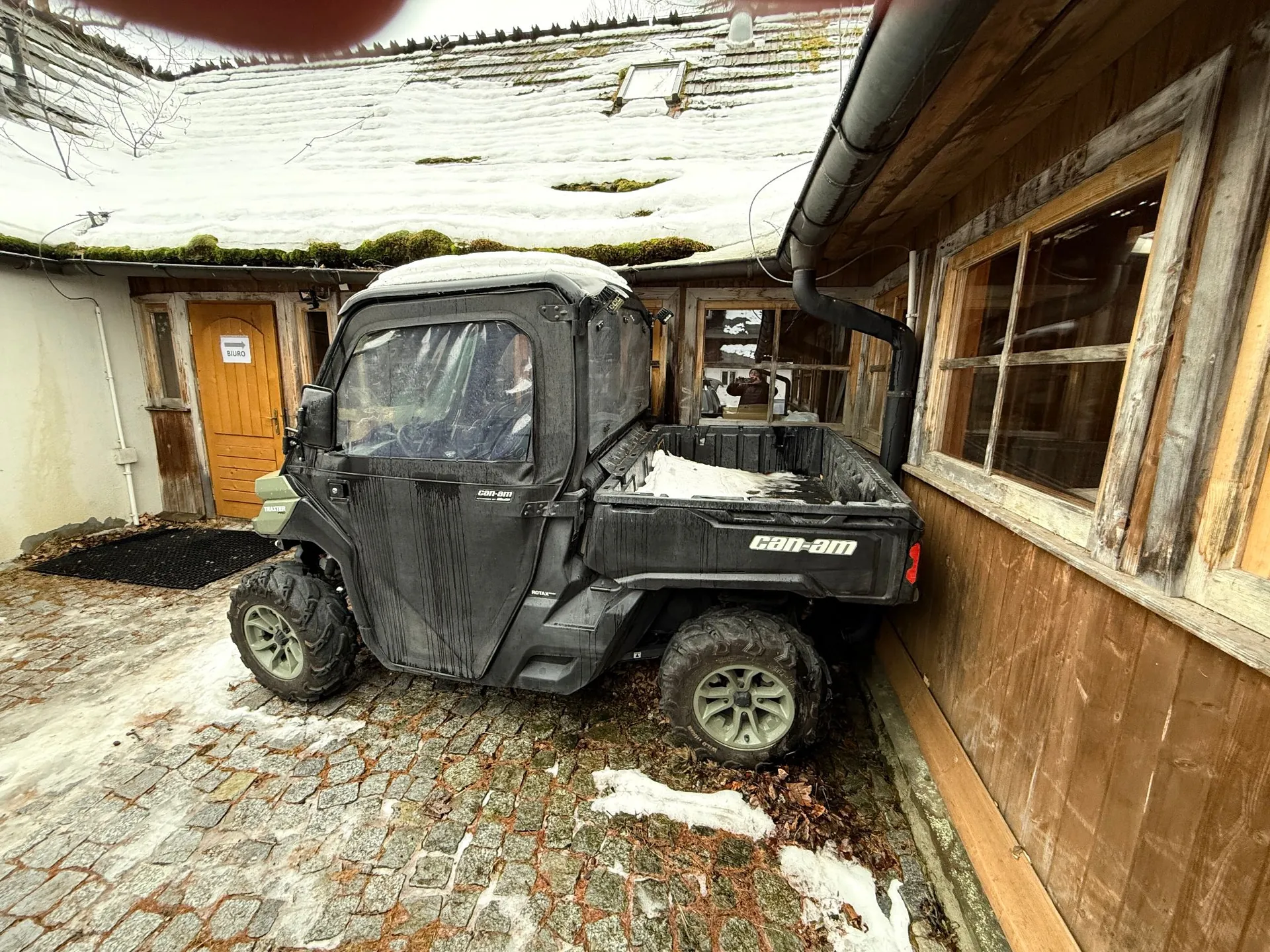 Czarny pojazd terenowy marki Can-Am z kabiną i otwartą paką, zaparkowany na brukowanym podjeździe przed drewnianym budynkiem. Na dachu budynku oraz na ziemi zalega śnieg. Na drewnianych drzwiach w tle widnieje biała tabliczka z napisem „BIURO”.