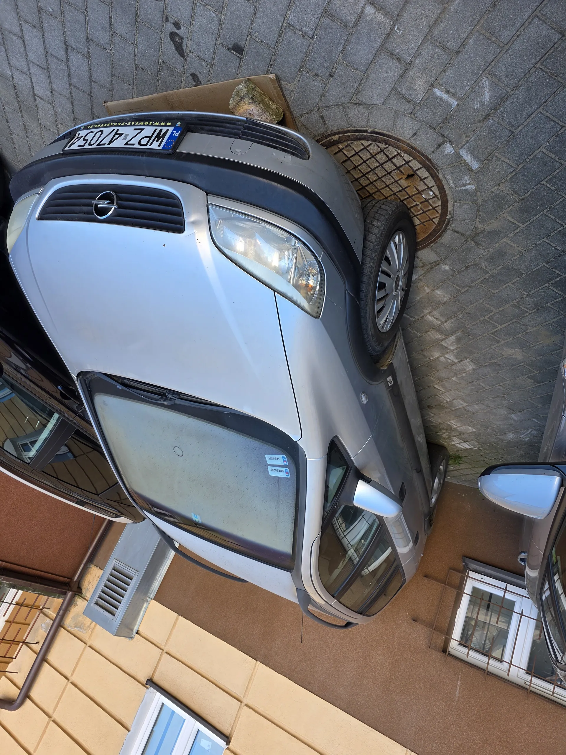 Srebrny samochód marki Opel zaparkowany na brukowanej nawierzchni. Widoczna tablica rejestracyjna WPZ 47054. W tle budynek z żółto-brązową elewacją, oknami z kratami i wentylatorem. Częściowo widoczny inny ciemny samochód. Przy przednim kołem srebrnego Opela kanałowa pokrywa, a obok niego karton z kamieniem. Na przedniej szybie samochodu naklejki.