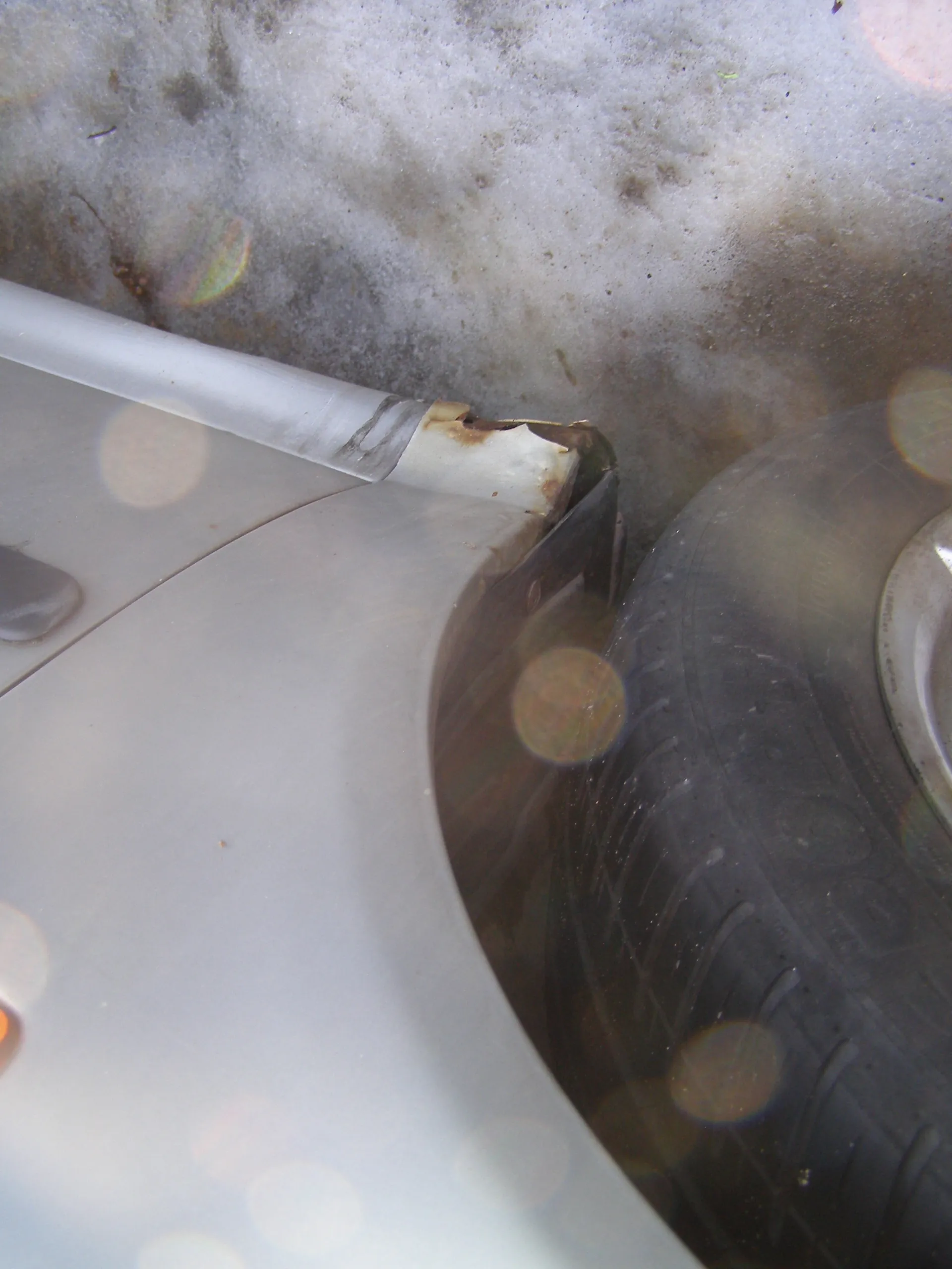 Zbliżenie na srebrny samochód, widoczna część nadkola z uszkodzonym białym elementem plastikowym z rdzą, opona z widocznymi śladami, zasneżona i zabrudzona powierzchnia ziemi oraz efekty świetlne w postaci plam od światła słonecznego.