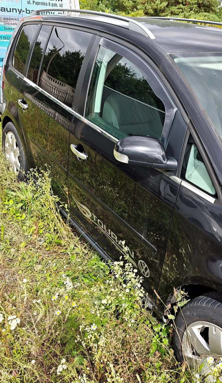 Zbliżenie na prawy bok czarnego samochodu typu kombi, zaparkowanego w wysokiej trawie z białymi polnymi kwiatami. Na dolnej części drzwi widoczny jest napis 'TOURER' oraz logo marki Volkswagen. W tle widoczny jest fragment białego znaku z tekstem 'ul. Paprolna 8 pok. 40'.