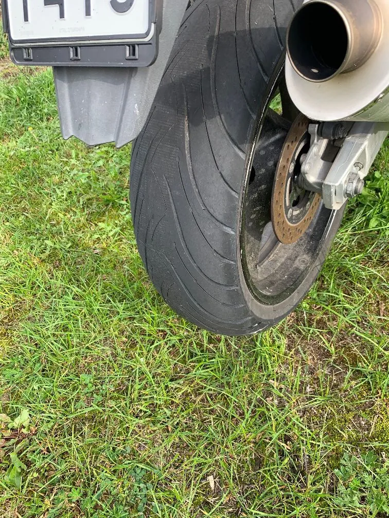 Zbliżenie na tylną oponę motocykla stojącego na zielonej trawie. Widoczny jest bieżnik opony, tarcza hamulcowa oraz fragment rury wydechowej i tablicy rejestracyjnej w górnej części obrazu.