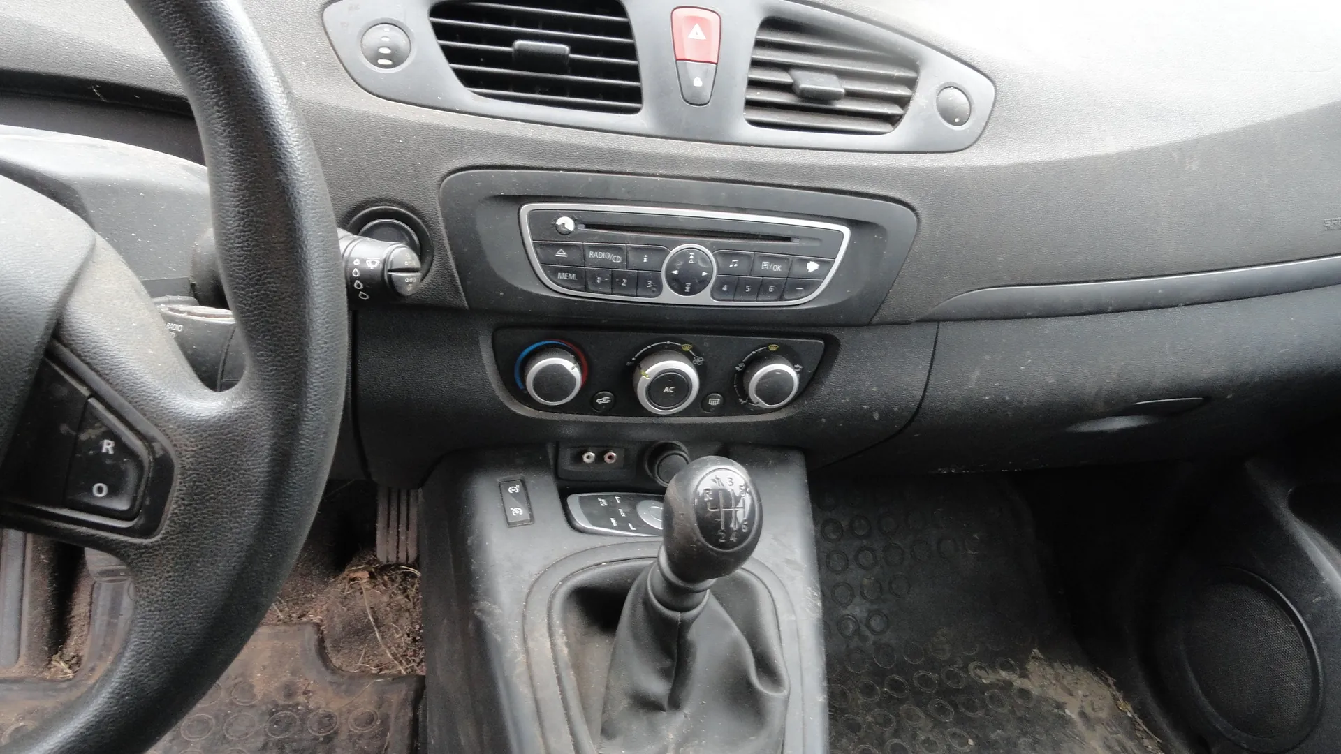 Wnętrze samochodu z widocznym czarnym kierownicą po lewej stronie, na której znajdują się przyciski oznaczone literami R i O. W centralnej części deski rozdzielczej umieszczono radio/CD z przyciskami MEM, 1-6, przyciskiem AC oraz trzema okrągłymi regulatorami klimatyzacji. Poniżej znajduje się dźwignia zmiany biegów z oznaczeniem 6-biegowej skrzyni biegów. Deska rozdzielcza i konsola środkowa są ciemnoszare, a dywaniki podłogowe brudne z widocznymi śladami ziemi. W górnej części deski rozdzielczej widoczne są wywietrzniki i czerwony przycisk ostrzegawczy.