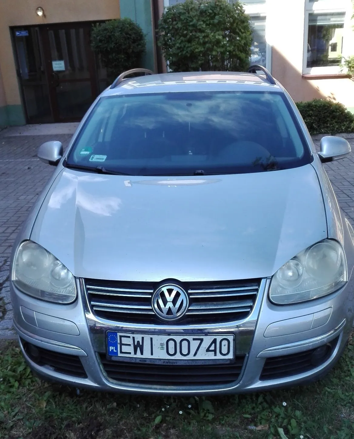 Srebrny samochód marki Volkswagen zaparkowany przed budynkiem. Widoczny jest przód pojazdu z logo producenta na grillu oraz polska tablica rejestracyjna o numerze EWI 00740. W tle widać wejście do budynku z ciemnymi drzwiami oraz zielone krzewy.