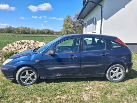Niebieski samochód osobowy typu hatchback zaparkowany na trawie obok białego budynku. Po lewej stronie widoczna jest sterta ziemi lub siana, a w tle rozległe pole i drzewa pod błękitnym niebem z kilkoma chmurami. Samochód posiada srebrne felgi, widoczne ślady korozji na boku oraz czerwony tylny reflektor. Budynek ma ciemny dach i białą elewację z czarną listwą na dole.
