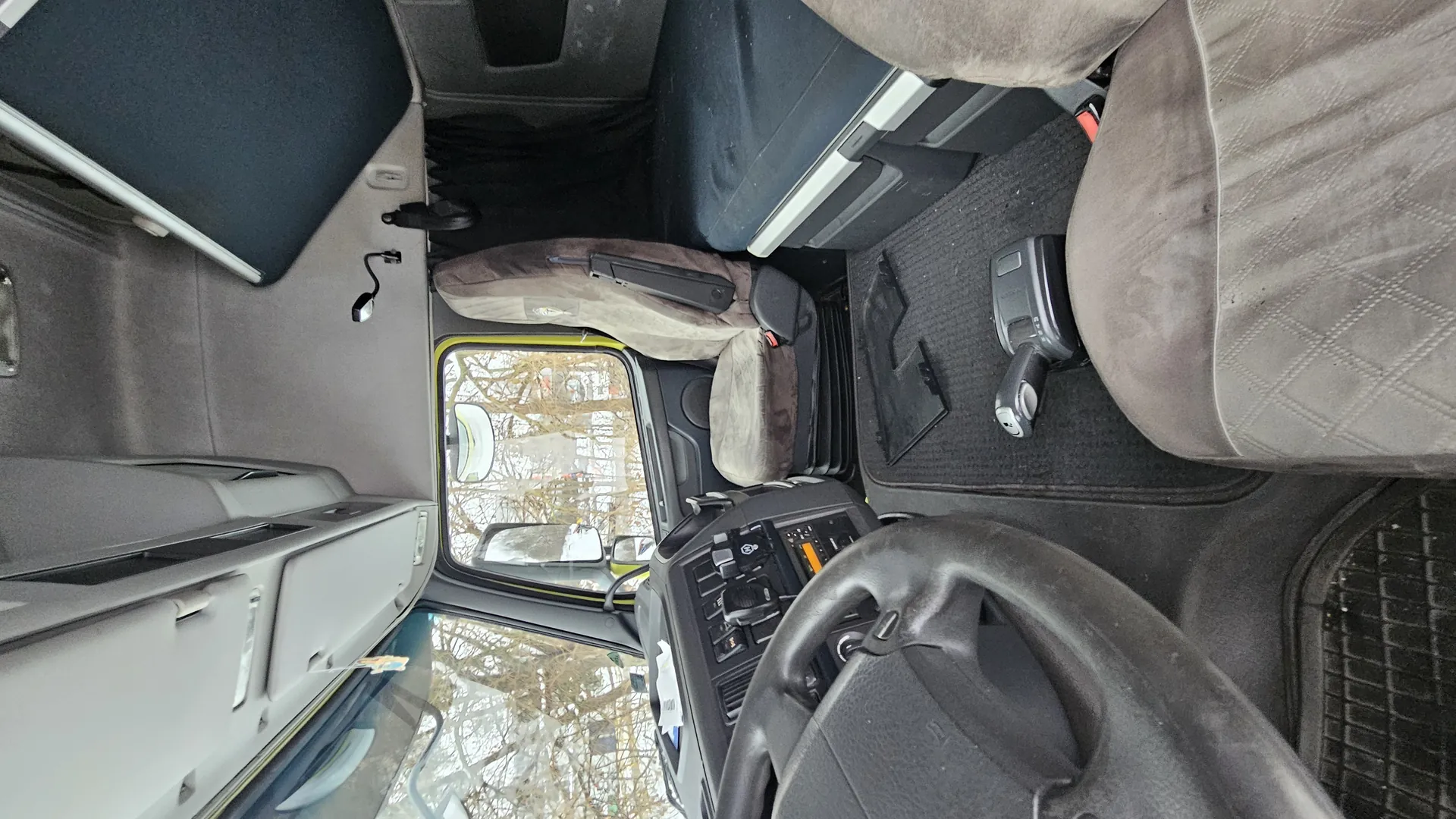 Wnętrze kabiny ciężarówki z kierownicą, deską rozdzielczą z przyciskami, szarymi siedzeniami i oknem pokazującym drzewa bez liści. Na podłodze czarny dywanik, odkurzacz oraz elementy wyposażenia. Brak czytelnego tekstu.