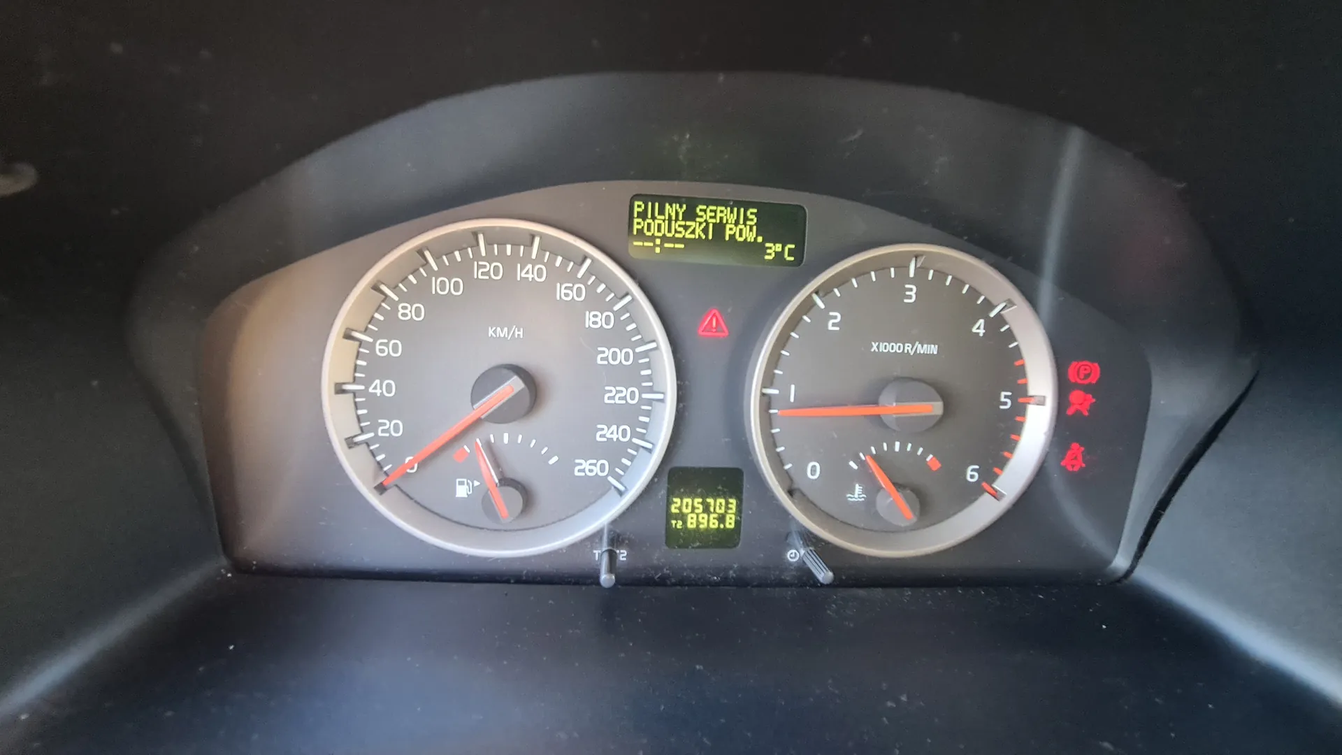 Tablica rozdzielcza samochodu z dwoma głównymi wskaźnikami. Po lewej stronie wskaźnik prędkości w km/h (0-260) z czerwoną igłą na 0 i pod nim wskaźnikiem paliwa. Po prawej stronie tachometr (0-6 x1000 R/MIN) z czerwoną igłą na 0 i pod nim wskaźnikiem temperatury chłodniczki. W centralnej części dwa wyświetlacze cyfrowe: górny z napisem 'PILNY SERWIS PODUSZKI POW. 3°C', dolny z liczbami '205703' i '896.8'. Widoczne czerwone lampki ostrzegawcze: trójkąt, pas bezpieczeństwa i inne symbole. Tło tablicy rozdzielczej jest ciemnoszare.