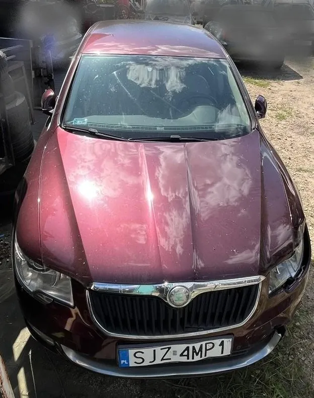 Widok z przodu na bordowy samochód osobowy marki Škoda. Pojazd posiada srebrną ramkę grilla z logo marki w centrum oraz tablicę rejestracyjną o numerze SJZ 4MP1. Na masce i przedniej szybie widoczne są odbicia chmur.