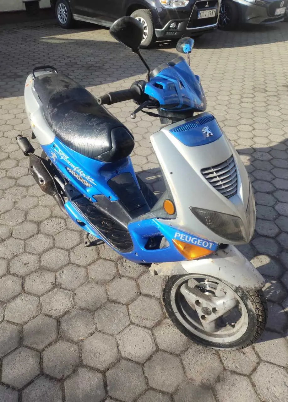 Niebiesko-srebrny skuter marki Peugeot zaparkowany na chodniku z sześciokątnych płytek. Na przedniej części skutera widoczny napis 'PEUGEOT' oraz logo marki. W tle czarny i szary samochód osobowy z tablicą rejestracyjną 'CLI 11XX'. Skuter ma czarne siedzenie, lusterka boczne i przednią maskę z niebieskim akcentem. Oświetlenie słoneczne, cienie wskazują na dzień.