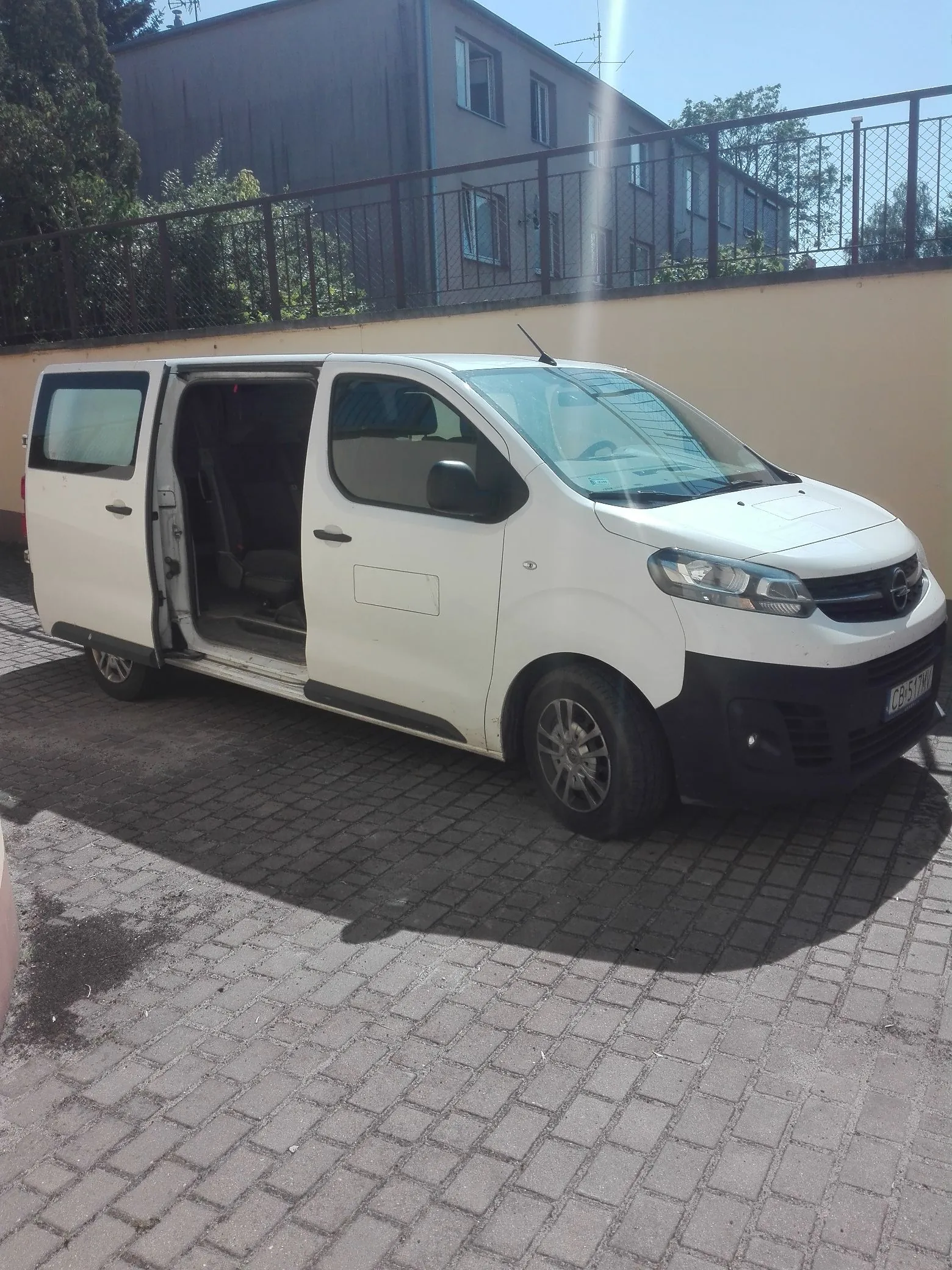 Biały samochód dostawczy marki Opel zaparkowany na kostce brukowej. Pojazd ma otwarte przesuwne drzwi boczne. W tle widoczny jest niski mur z metalowym ogrodzeniem oraz wielopiętrowy budynek mieszkalny pod jasnym, słonecznym niebem.