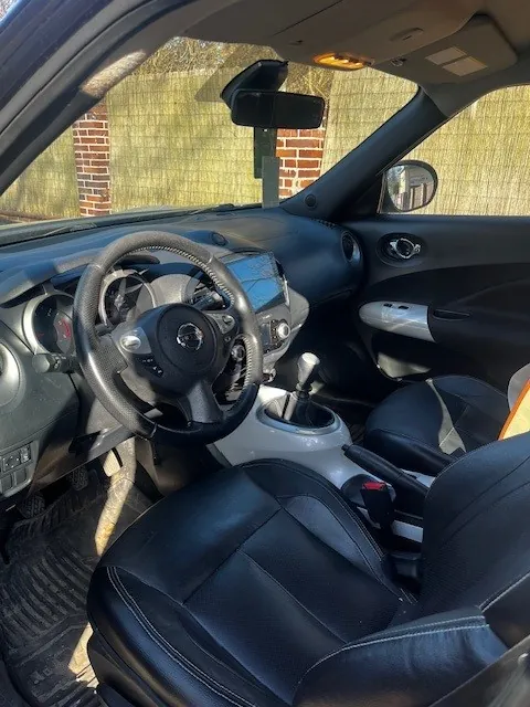 Wnętrze samochodu z czarnymi skórzonymi fotelami, kierownicą z logo, deską rozdzielczą z wskaźnikami, dźwignią zmiany biegów, lusterkiem wstecznym i bocznym. Przez przednią szybę widoczna ceglana ściana i zieleń.