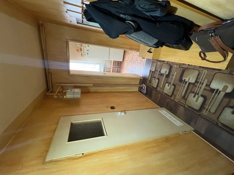 Wąski korytarz z drewnianymi ścianami. Po lewej stronie jasne drzwi z małym kwadratowym okienkiem, na podłodze dywan z geometrycznym wzorem. Przy drzwiach para fioletowych klap. Po prawej drewniana ławka z brązową torbą i ciemnymi kurtkami wiszącymi na haczkach. W głębi otwarte drzwi prowadzące do kuchni z białą lodówką i oknem. Na suficie widoczne rury i oświetlenie.