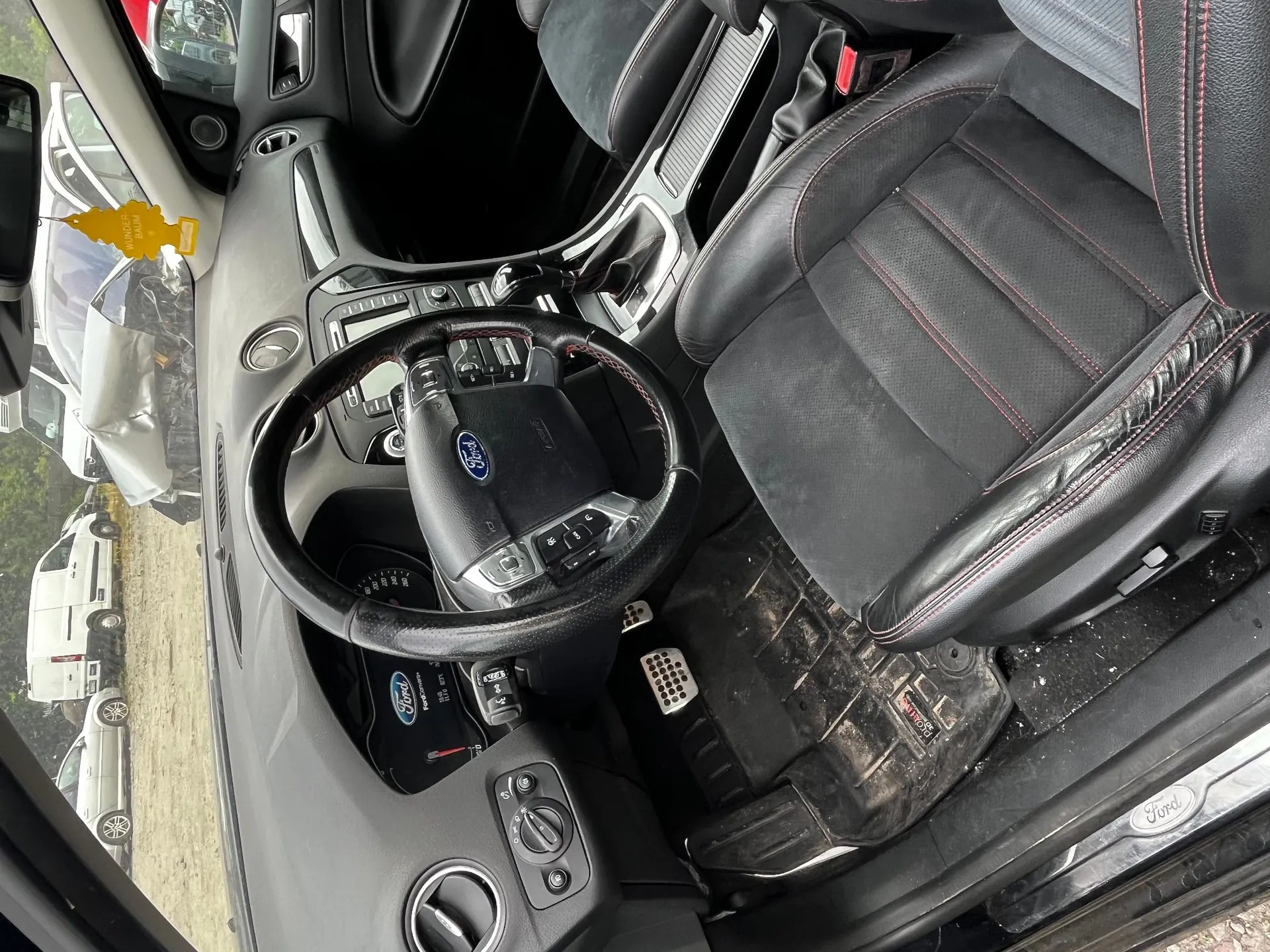 Wnętrze samochodu marki Ford, widok z perspektywy kierowcy. Widoczna czarna, skórzana kierownica z logo Ford, czarny fotel z czerwonymi przeszyciami, deska rozdzielcza oraz gumowa mata podłogowa. Przez przednią szybę i otwarte drzwi widoczne są inne uszkodzone pojazdy na placu. Na progu samochodu znajduje się srebrna tabliczka z napisem Ford.
