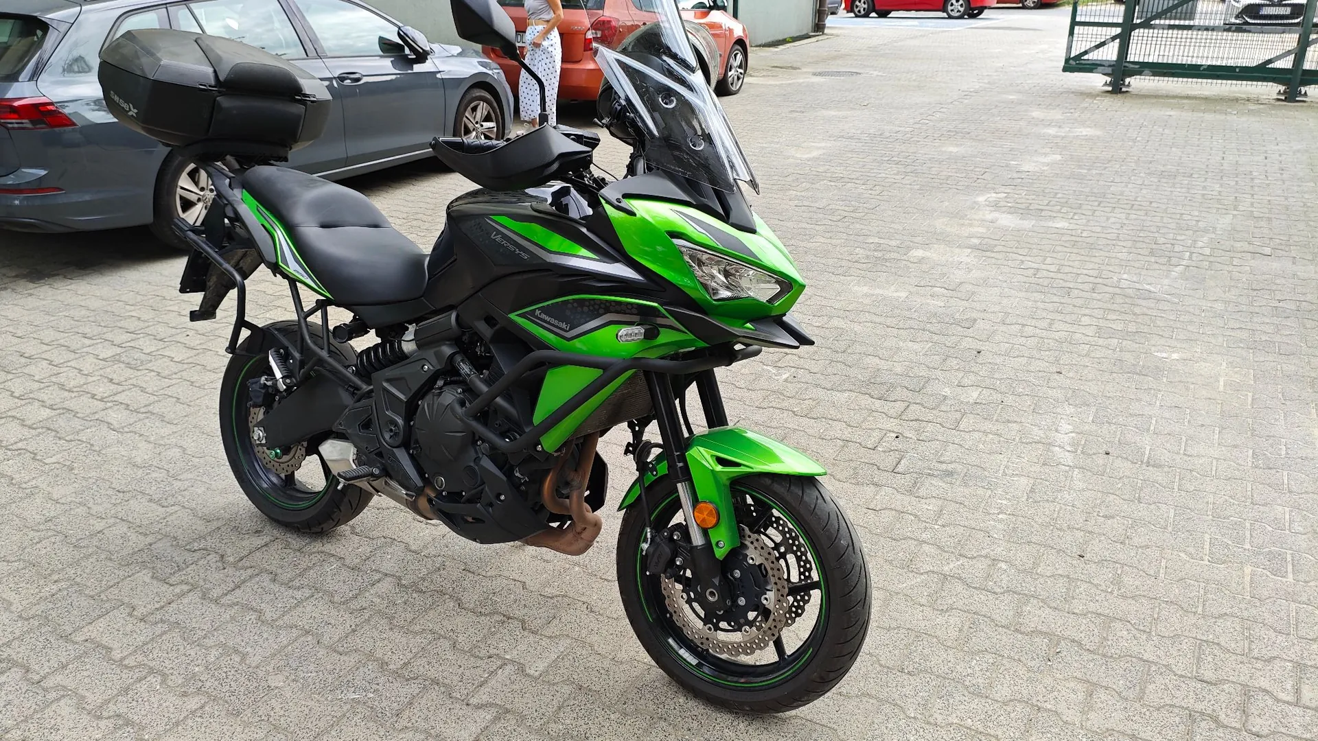Zielono-czarny motocykl Kawasaki Versys zaparkowany na kostce brukowej. Motocykl posiada czarny kufer z tyłu, wysoką szybę ochronną oraz zielone detale na nadwoziu i felgach. W tle widoczne są zaparkowane samochody: szary i pomarańczowy.