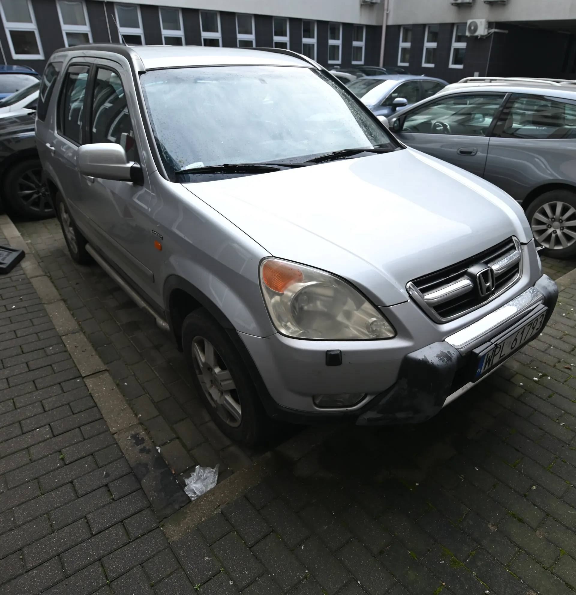 Srebrny samochód osobowy marki Honda typu SUV zaparkowany na brukowanym placu. Widoczna tablica rejestracyjna WPL 61793. W tle budynki z oknami, klimatyzatorami oraz inne zaparkowane pojazdy. Na nawierzchni leży plastikowy worek. Samochód ma czarny zderzak przedni i reflektory z pomarańczowymi kierunkowskazami.