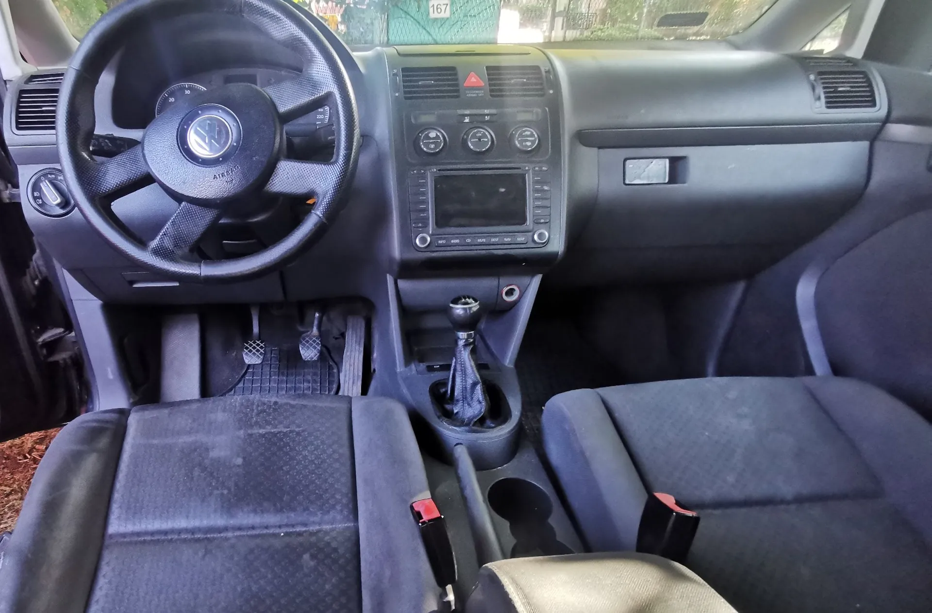 Wnętrze samochodu marki Volkswagen z perspektywy przednich siedzeń. Widoczna jest czarna kierownica z logo VW, deska rozdzielcza z systemem audio i klimatyzacją, lewarek zmiany biegów oraz dwa przednie fotele z ciemnej tkaniny.