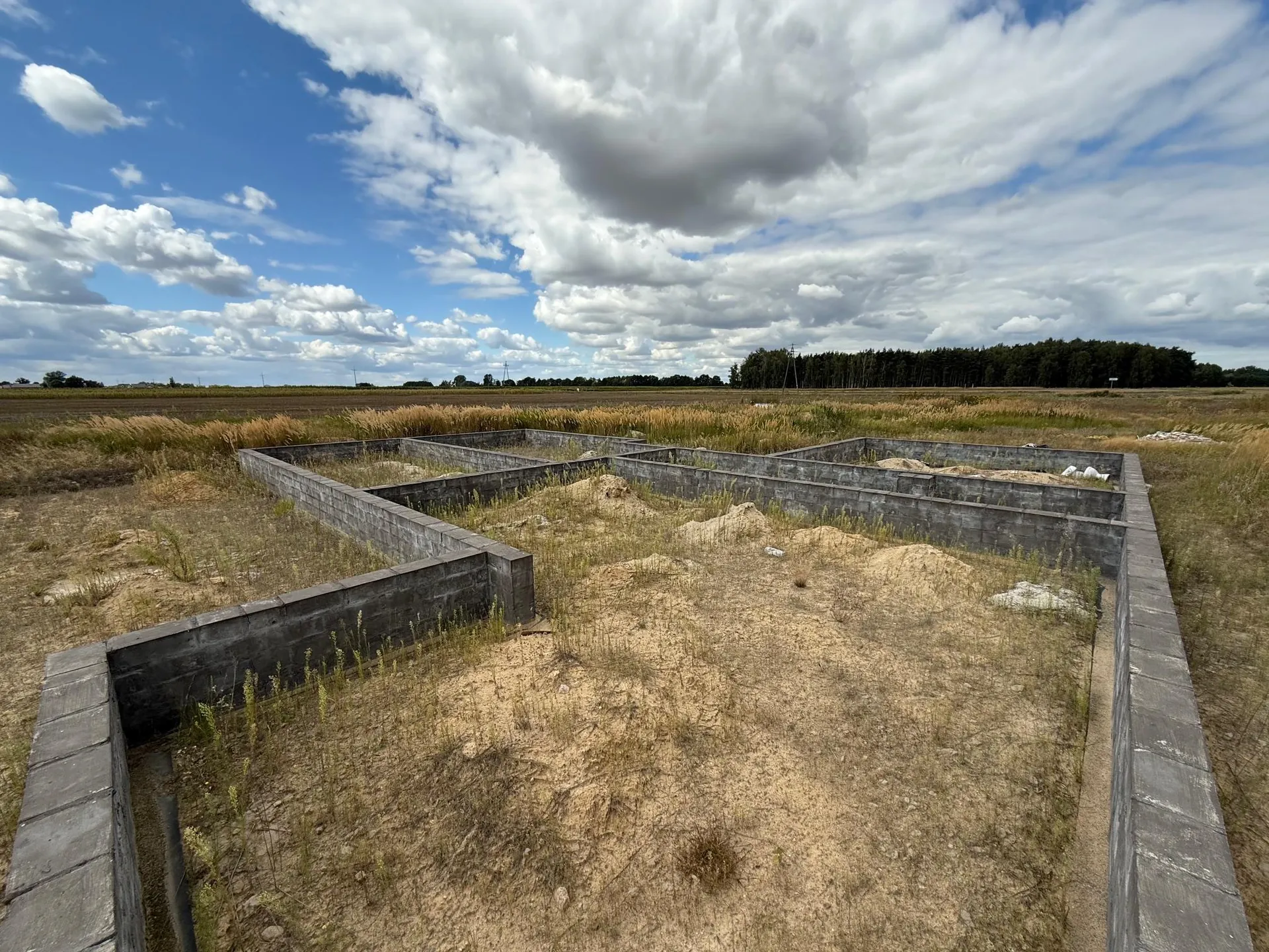 Zdjęcie przedstawiające fundamenty budynku w budowie, wykonane z szarych bloczków betonowych. Fundamenty tworzą kilka połączonych ze sobą prostokątnych pomieszczeń na piaszczystym terenie z kępami suchej trawy. W tle widać rozległe pole oraz linię gęstego lasu pod jasnoniebieskim niebem z licznymi białymi chmurami.