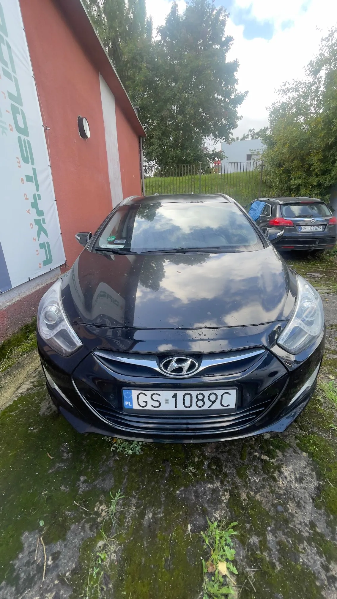 Czarny samochód marki Hyundai zaparkowany przodem do obiektywu na podłożu z mchu i betonu. Samochód ma polską tablicę rejestracyjną o numerze GS 1089C. W tle widoczny jest czerwony budynek z białym napisem 'LOGISTYKA medyczna', zielony płot oraz drzewa.