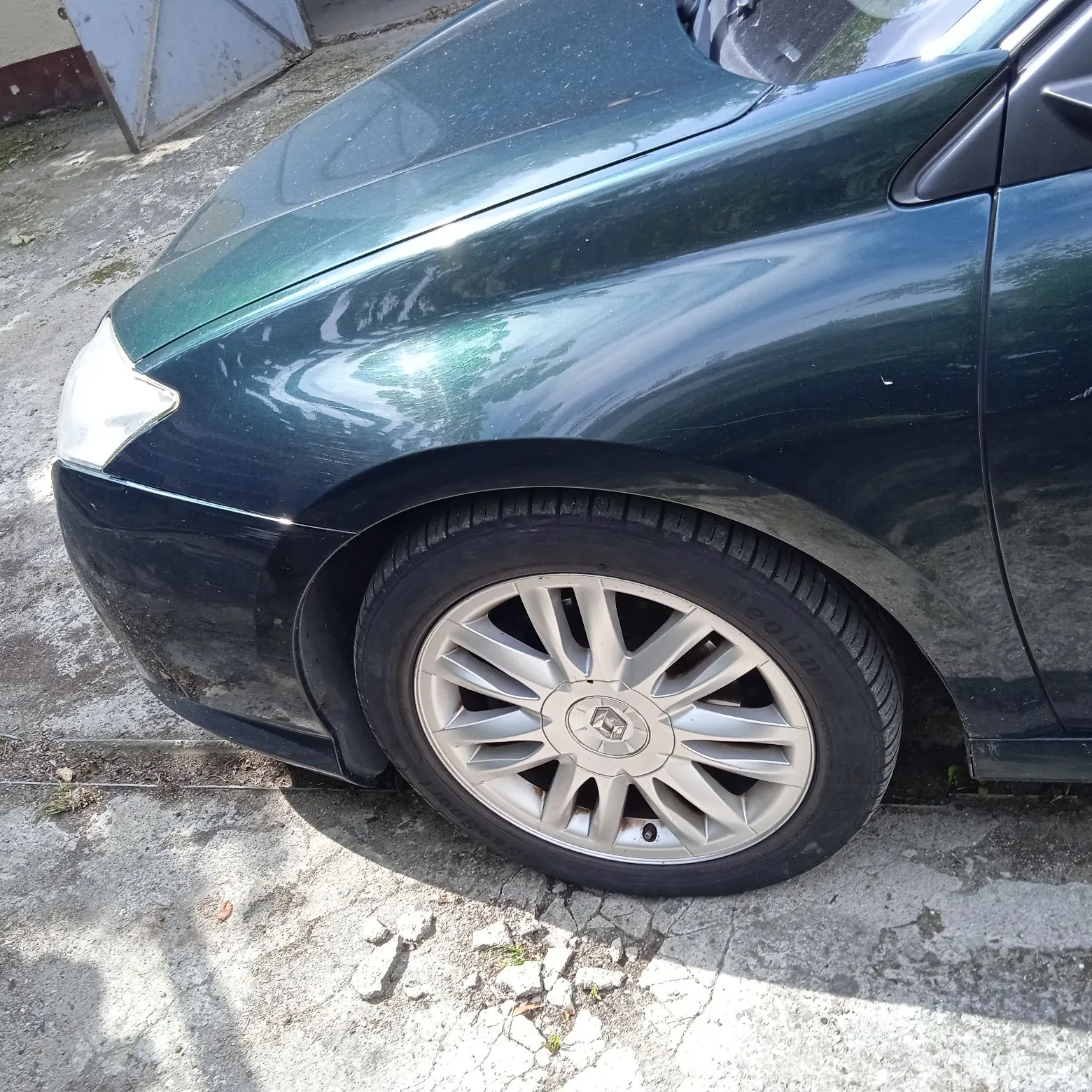Zbliżenie na przedni lewy błotnik i koło ciemnozielonego samochodu. Widoczna jest srebrna wieloramienna felga z logo producenta w centrum oraz opona marki Neolin. Samochód stoi na betonowym podłożu.